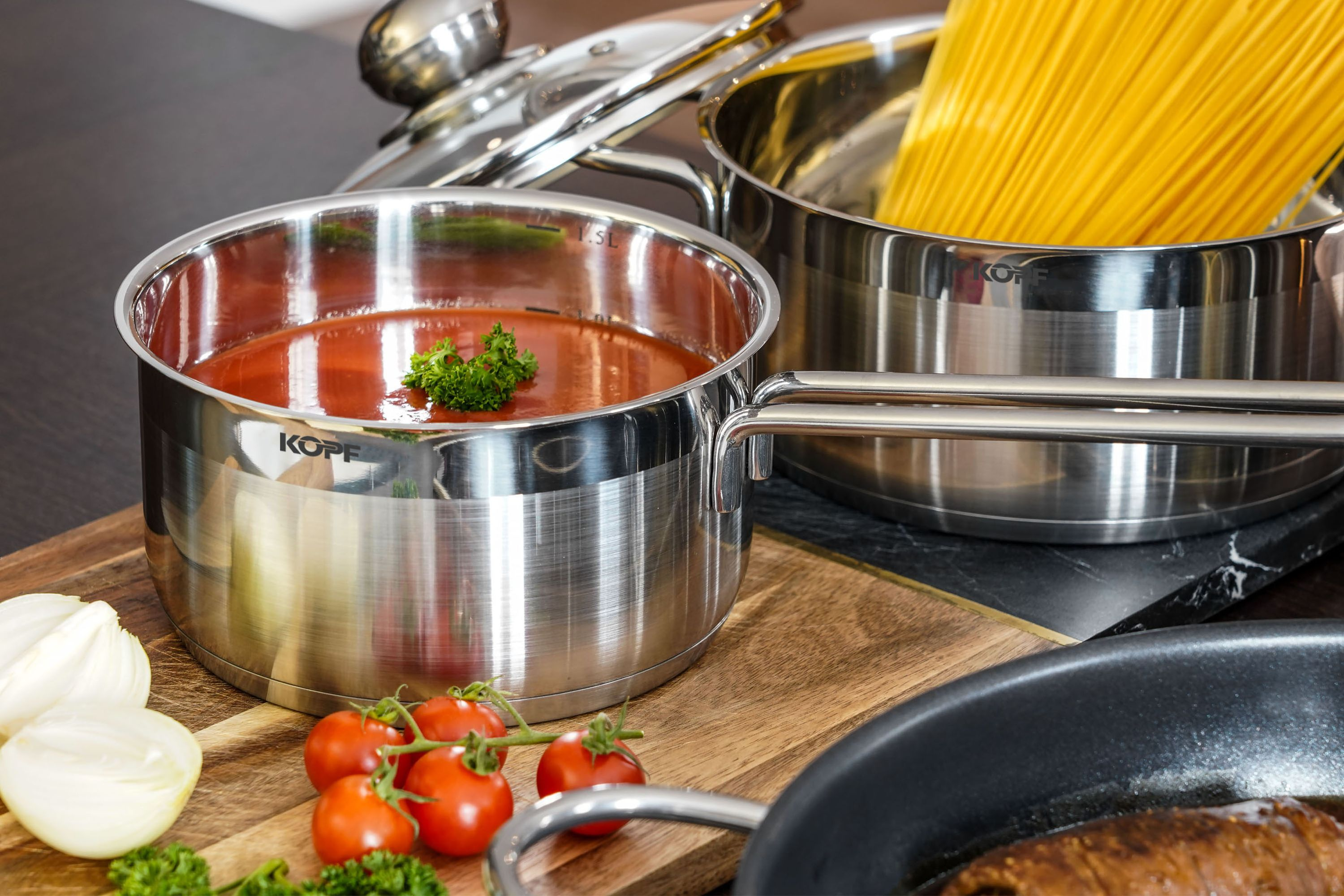 Edelstahltöpfe mit Spaghetti, Tomatensauce und Gemüse auf einem Holzschneidebrett, mit Tomaten und Zwiebeln.