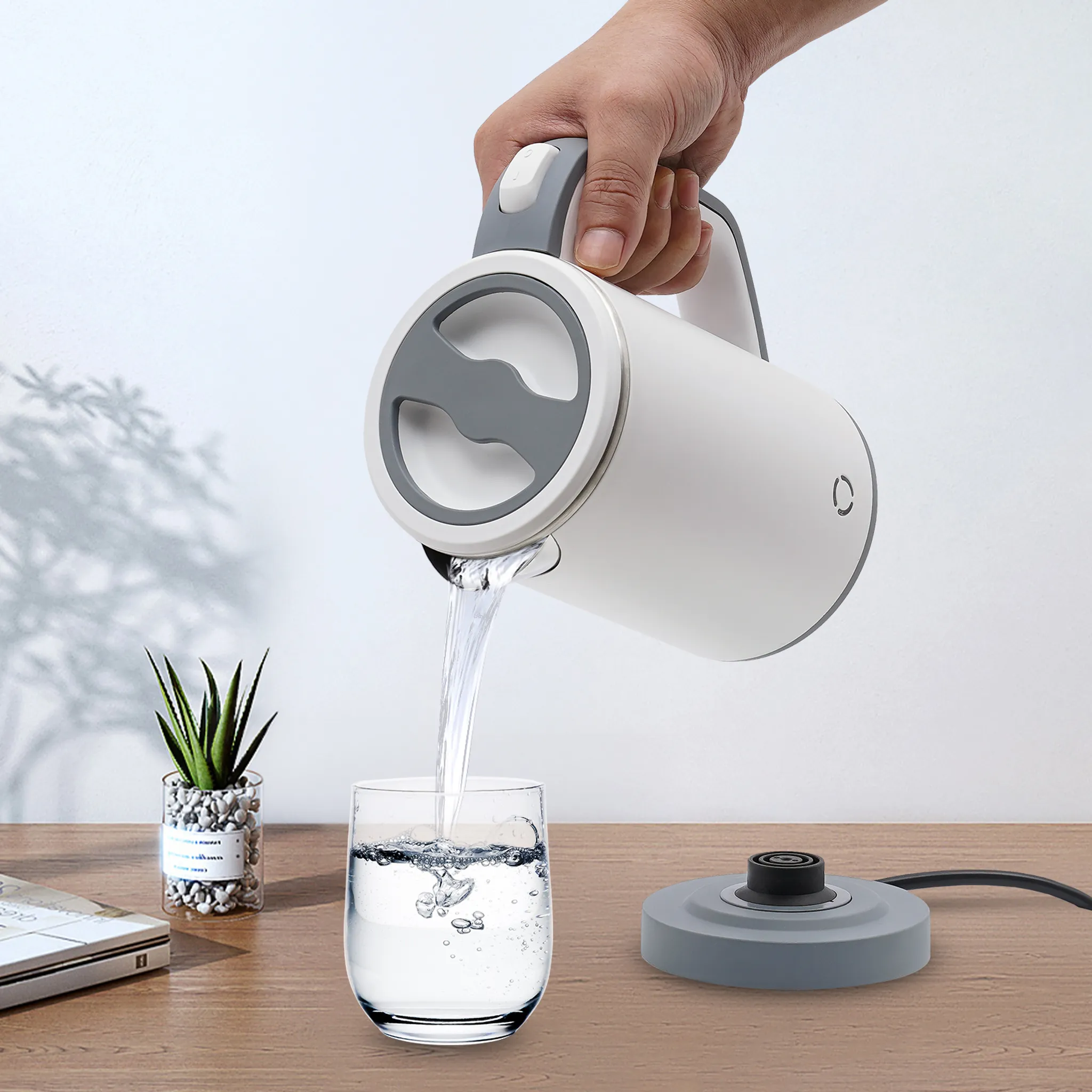 Eine Hand gießt Wasser aus einem weißen Wasserkocher in ein Glas. Auf einem Holztisch stehen eine Pflanze und ein Schalter.