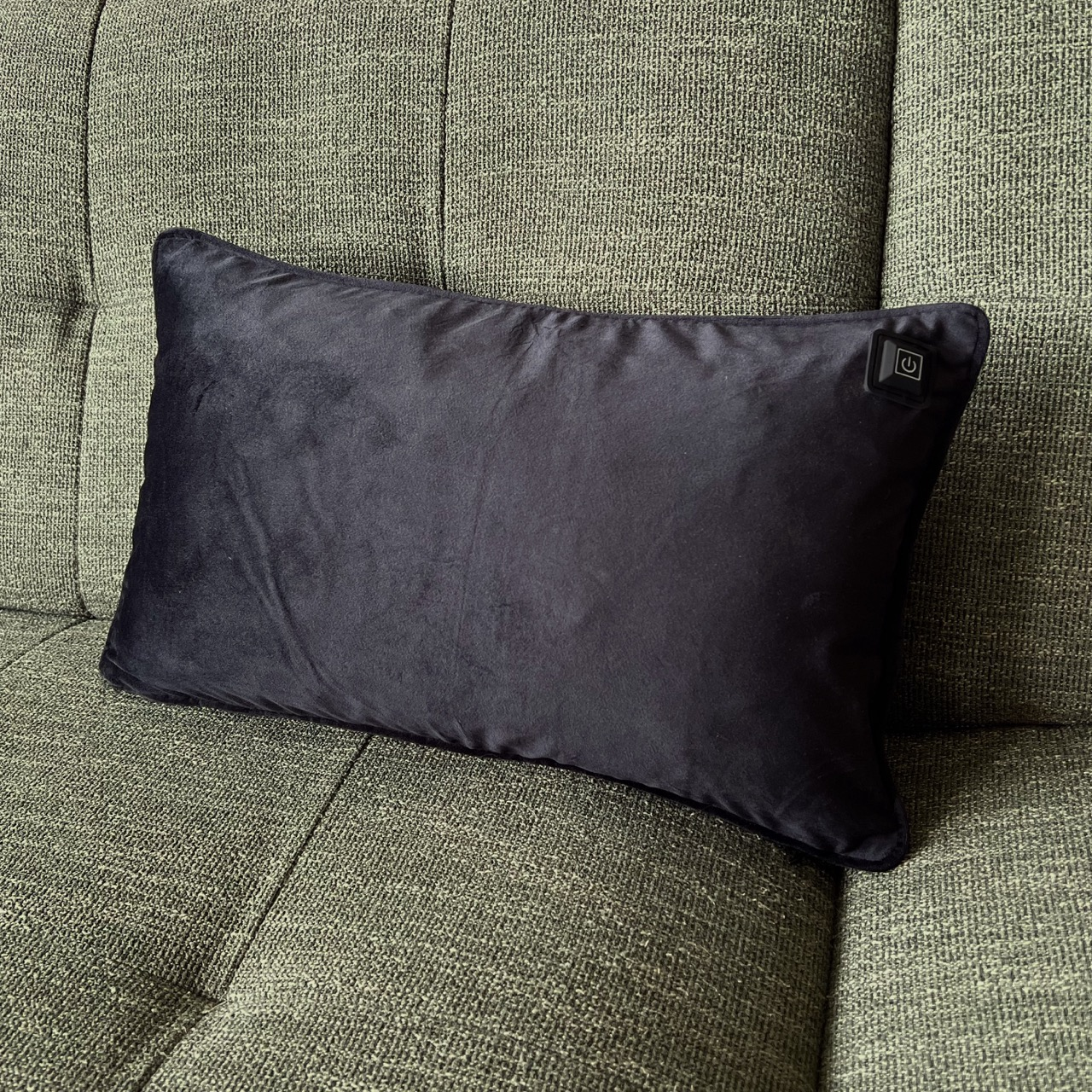 Ein schwarzes Kissen mit Power-Taste liegt auf einer grünen Couch. Das Kissen ist rechteckig und samtartig.