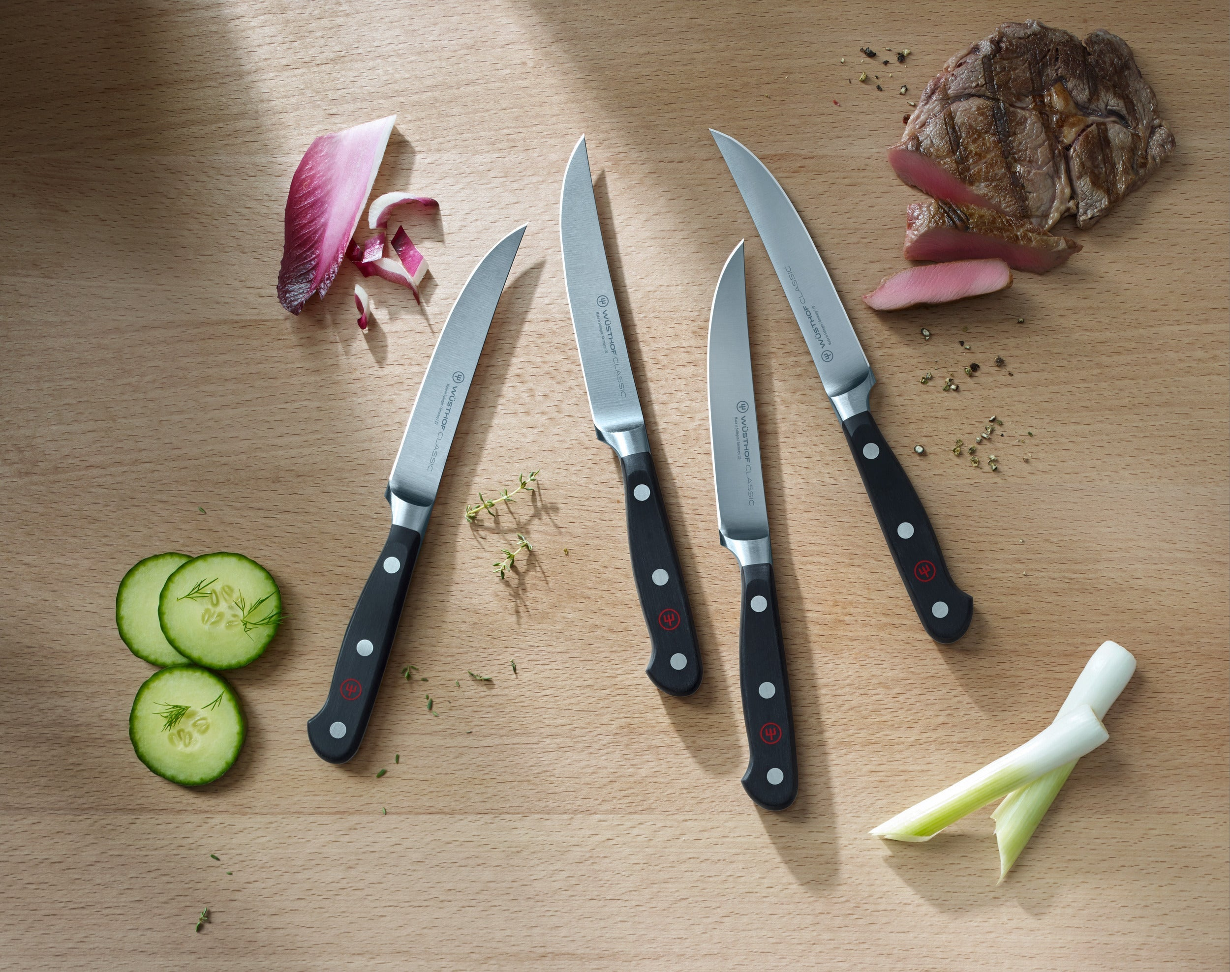 Vier Messer, Fleisch, Gurkenscheiben und roter Salat auf einem Holzbrett. Dill und Kräuter sind verstreut.