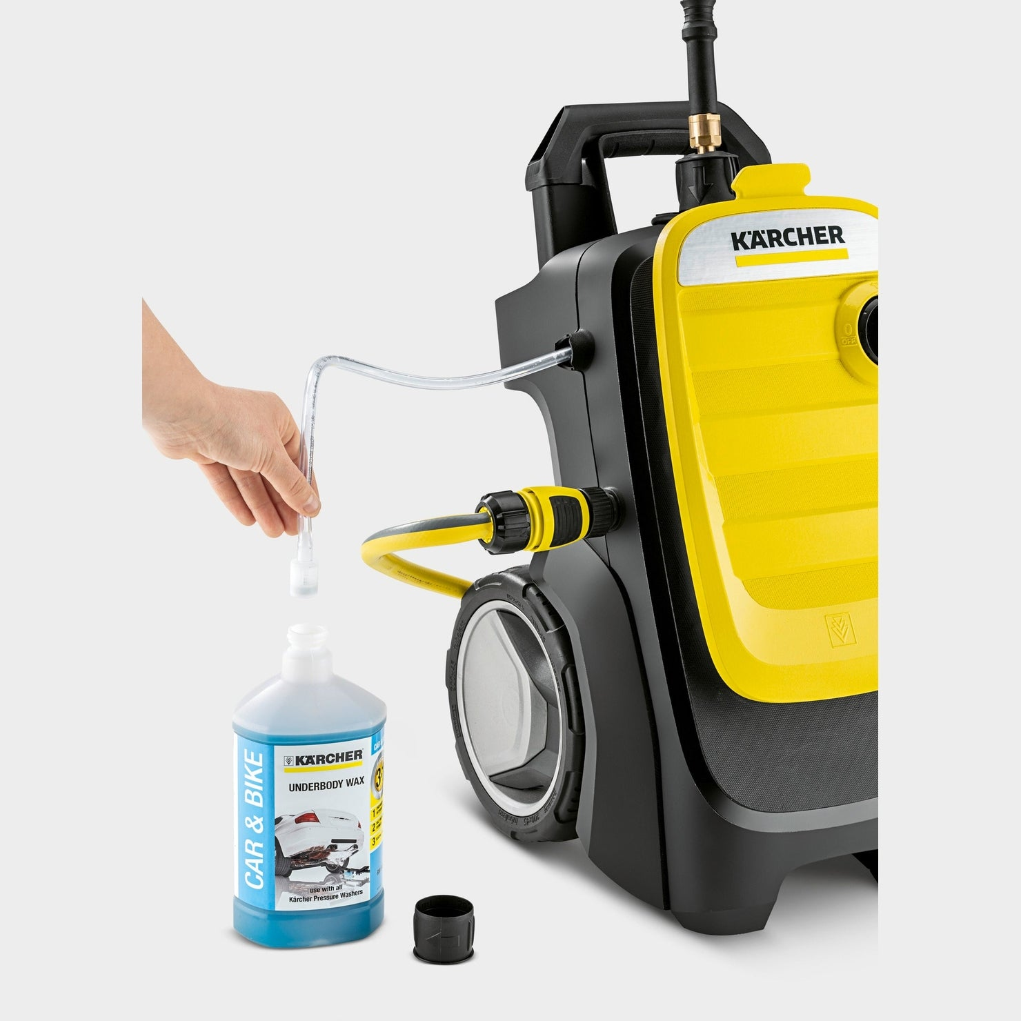 Una mano versa liquido da una bottiglia 'Kärcher Underbody Wax' in un'idropulitrice. L'idropulitrice è gialla e nera.