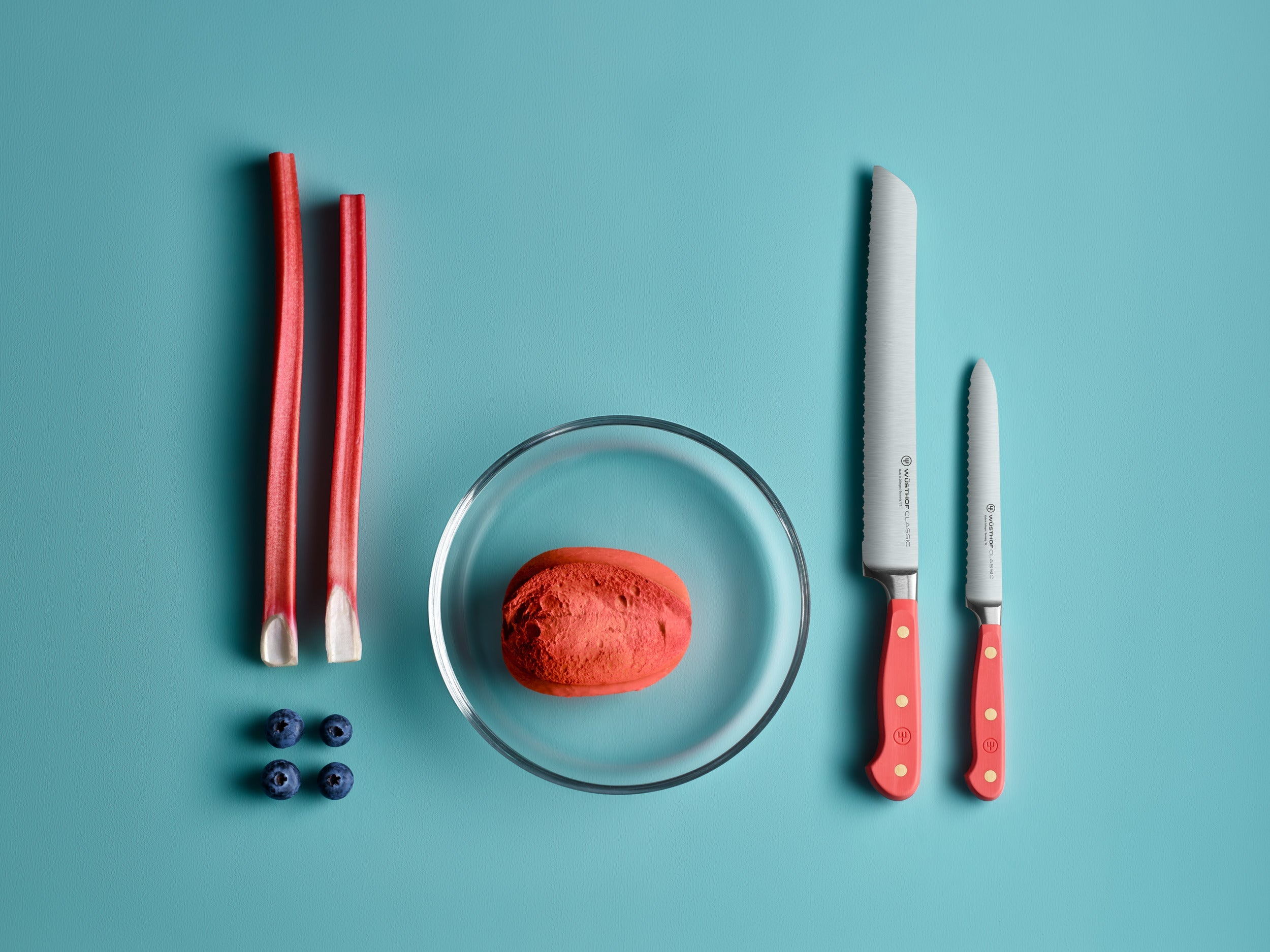 Rhabarberstiele, eine Glasschale mit rotem Inhalt, zwei Messer und Blaubeeren auf blauem Hintergrund.