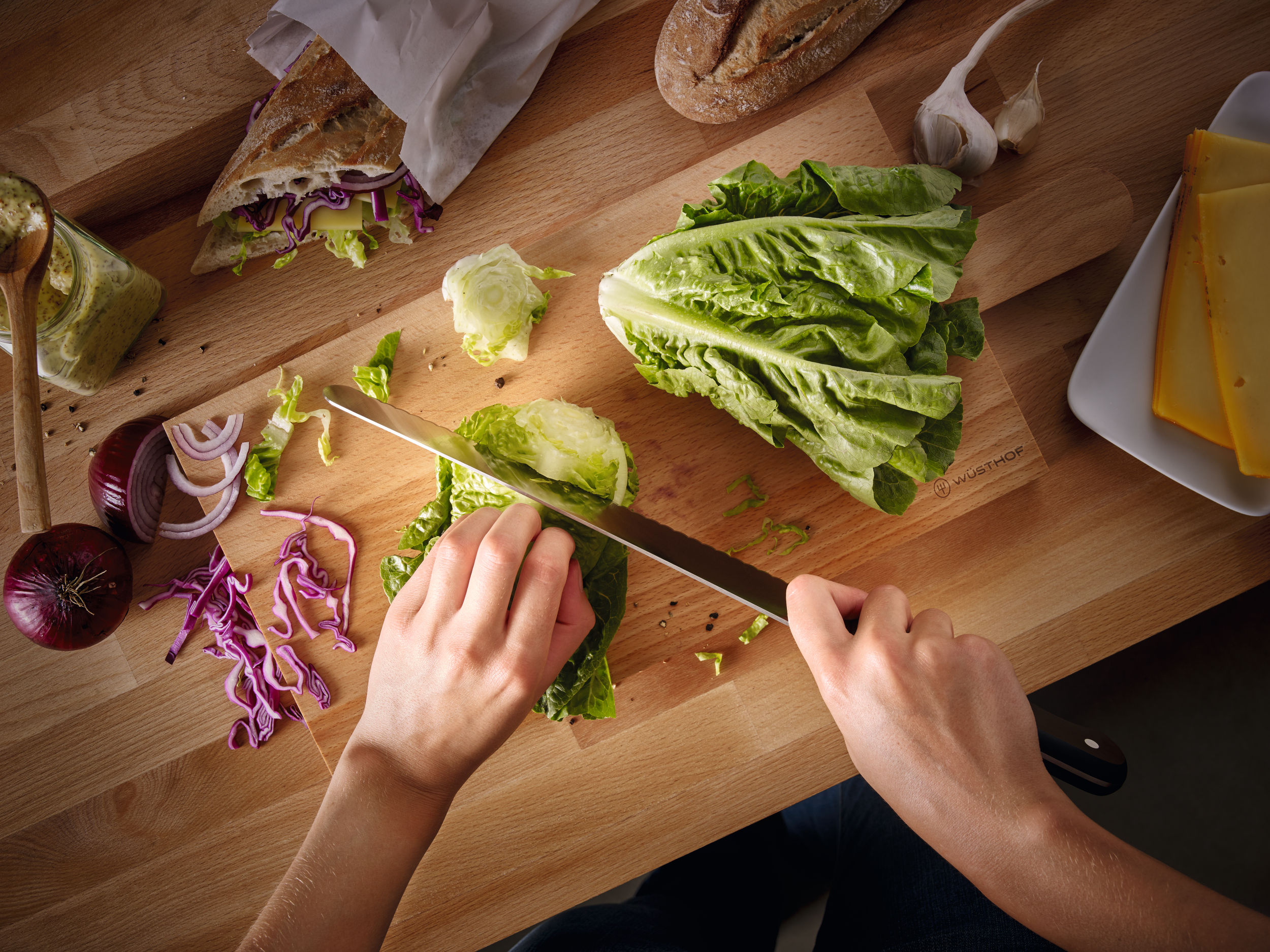 Hände schneiden Salat auf einem Holzschneidebrett. Andere Zutaten wie Käse, Brot und Zwiebeln sind vorhanden.