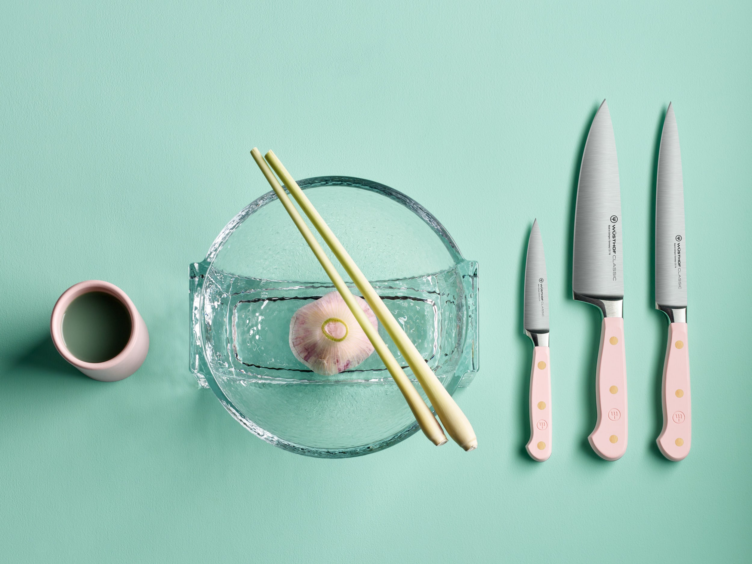 Ein Stillleben mit Wasser, Knoblauch, Zitronengras, einer Tasse und drei Messern mit rosa Griffen auf tealfarbenem Hintergrund.