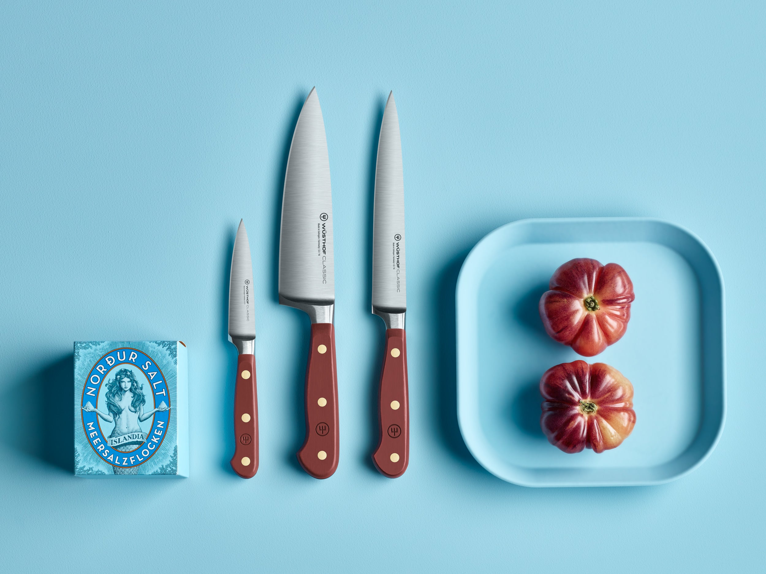 Tres cuchillos con mangos de madera, dos tomates en una bandeja azul y una caja de sal, todo sobre una superficie azul.