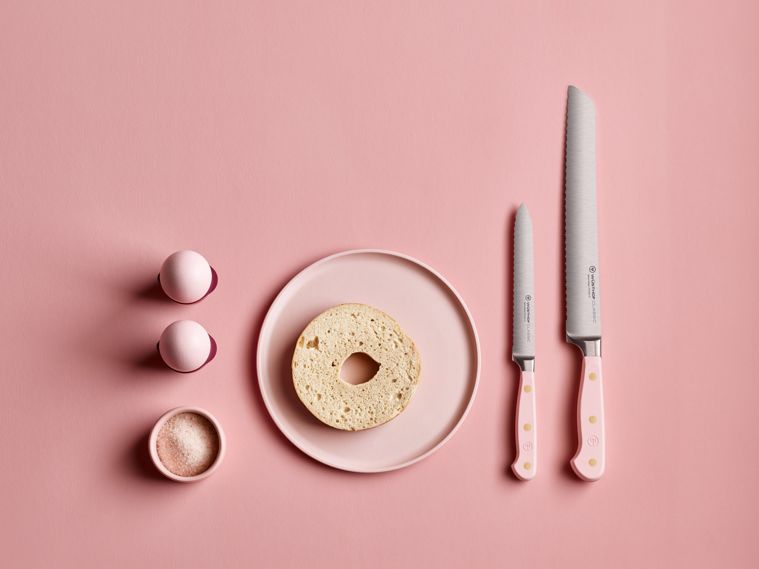 Rosa Hintergrund mit Bagel auf einem Teller, zwei Messern, zwei Eiern und einer Schüssel Salz. Alles ist rosa.