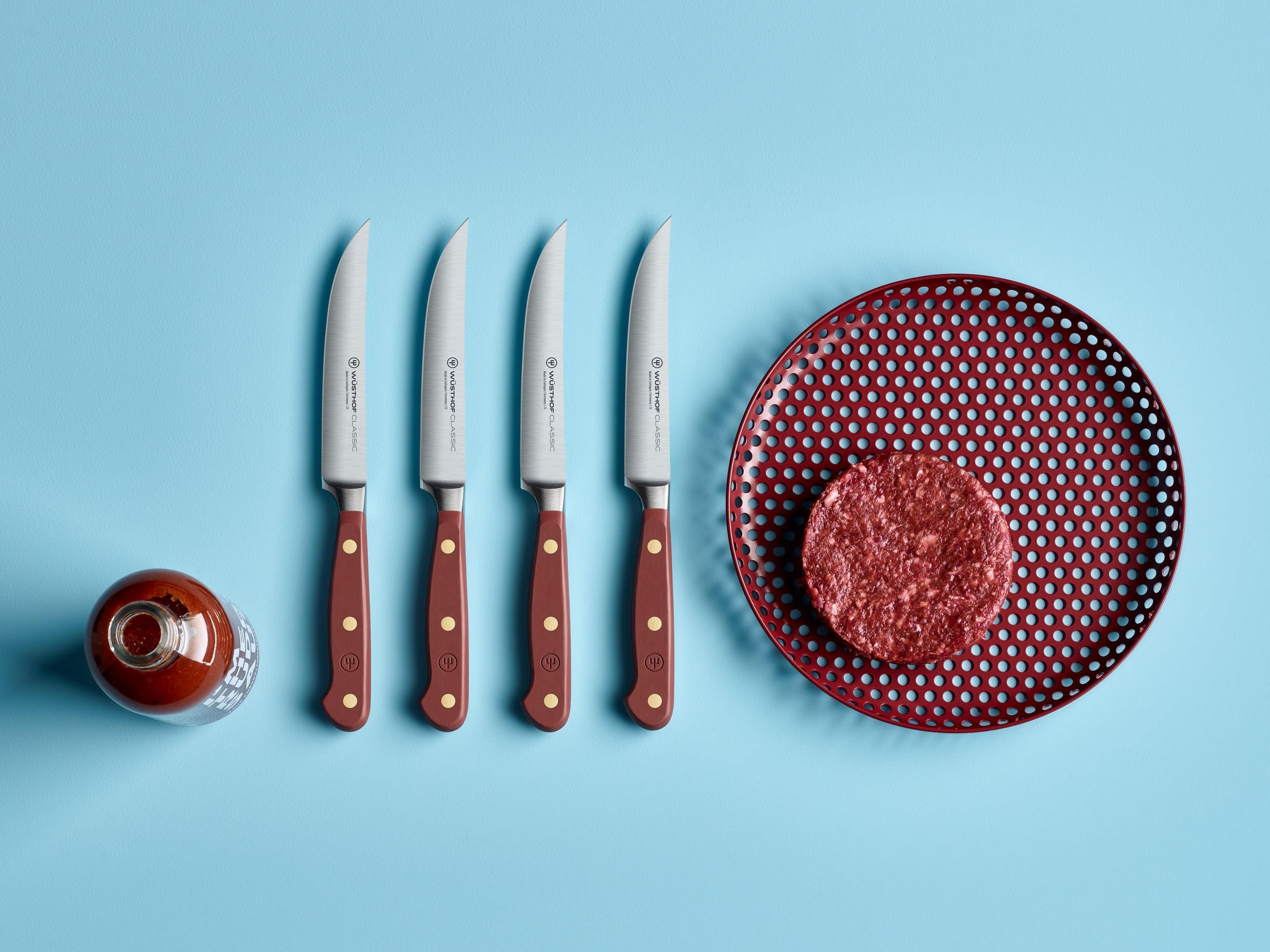 Vier Steakmesser, ein Burger-Patty auf einem roten Teller und eine Flasche Sauce auf hellblauem Grund.
