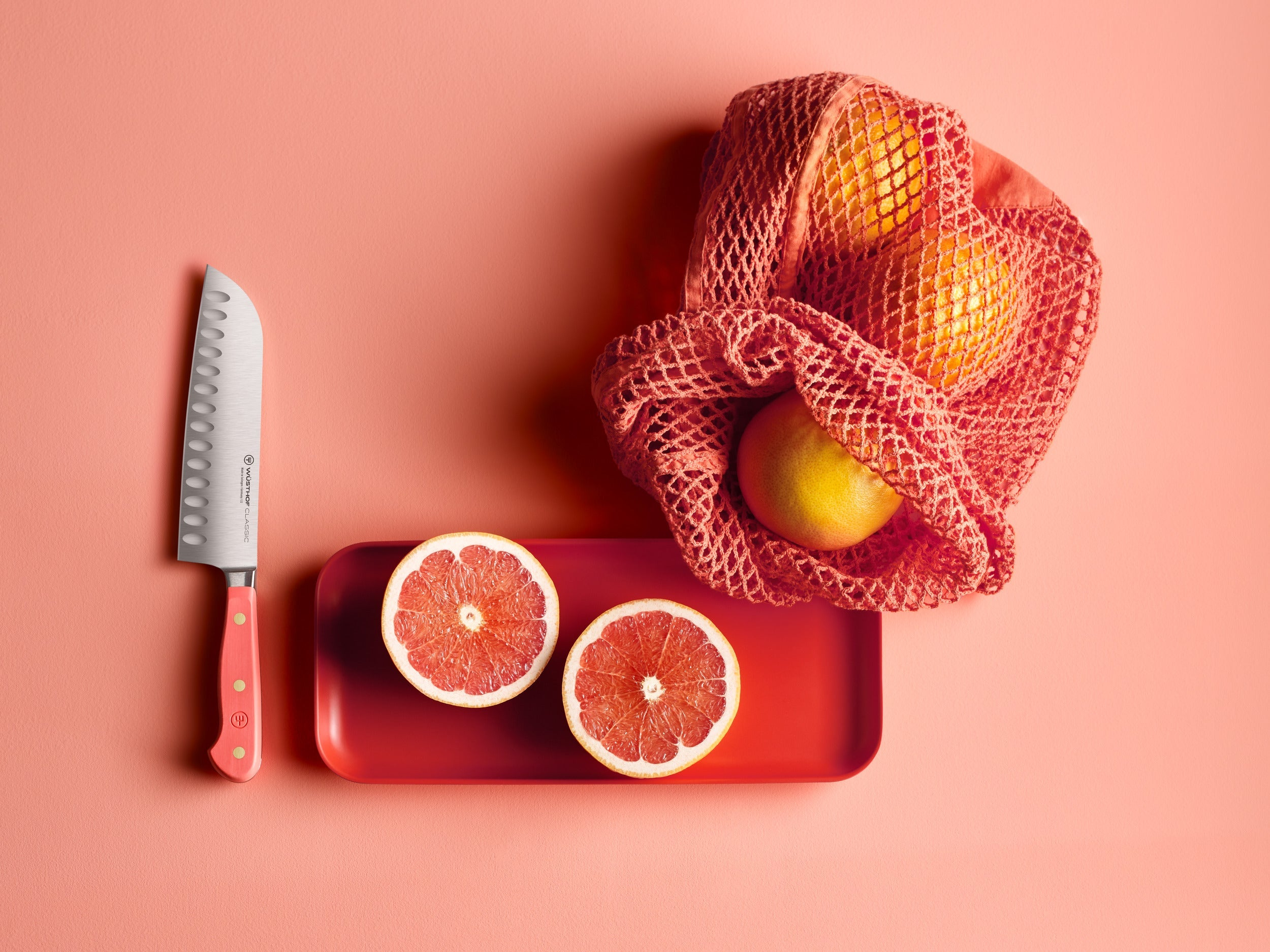 Ein Messer und aufgeschnittene Grapefruits auf einem roten Tablett. Orangen in einer roten Netztasche, alles auf pinkem Hintergrund.