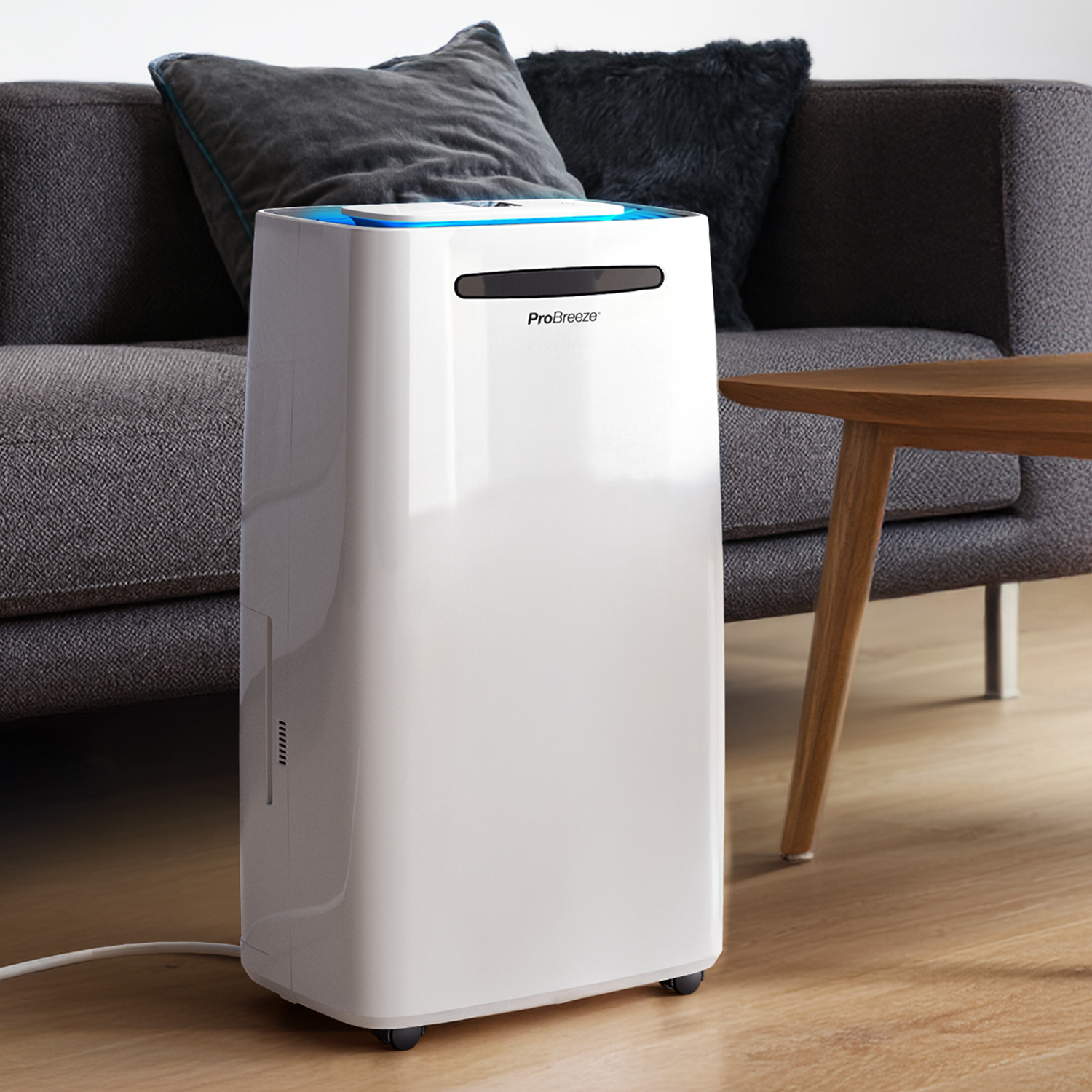 Weißer ProBreeze-Luftreiniger in einem Wohnzimmer. Graues Sofa und Kissen, ein Holztisch. Der Boden ist aus Holz.
