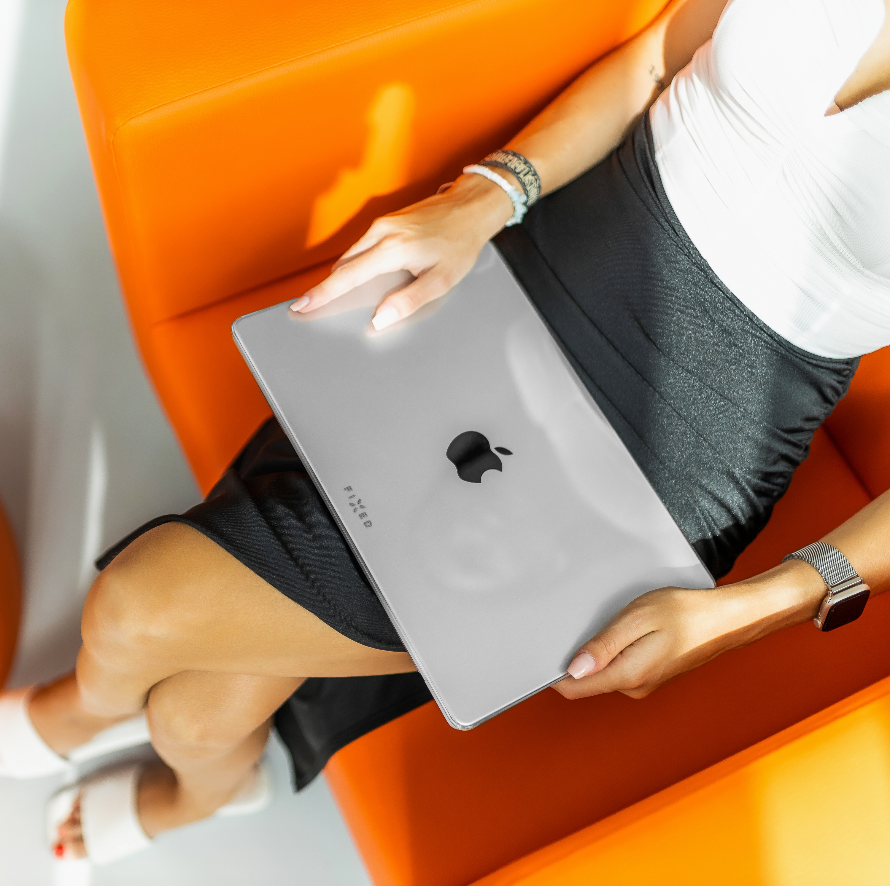 Frau mit Apple-Laptop auf dem Schoß, sitzt in einem orangefarbenen Stuhl. Sie trägt ein weißes Oberteil und einen schwarzen Rock.
