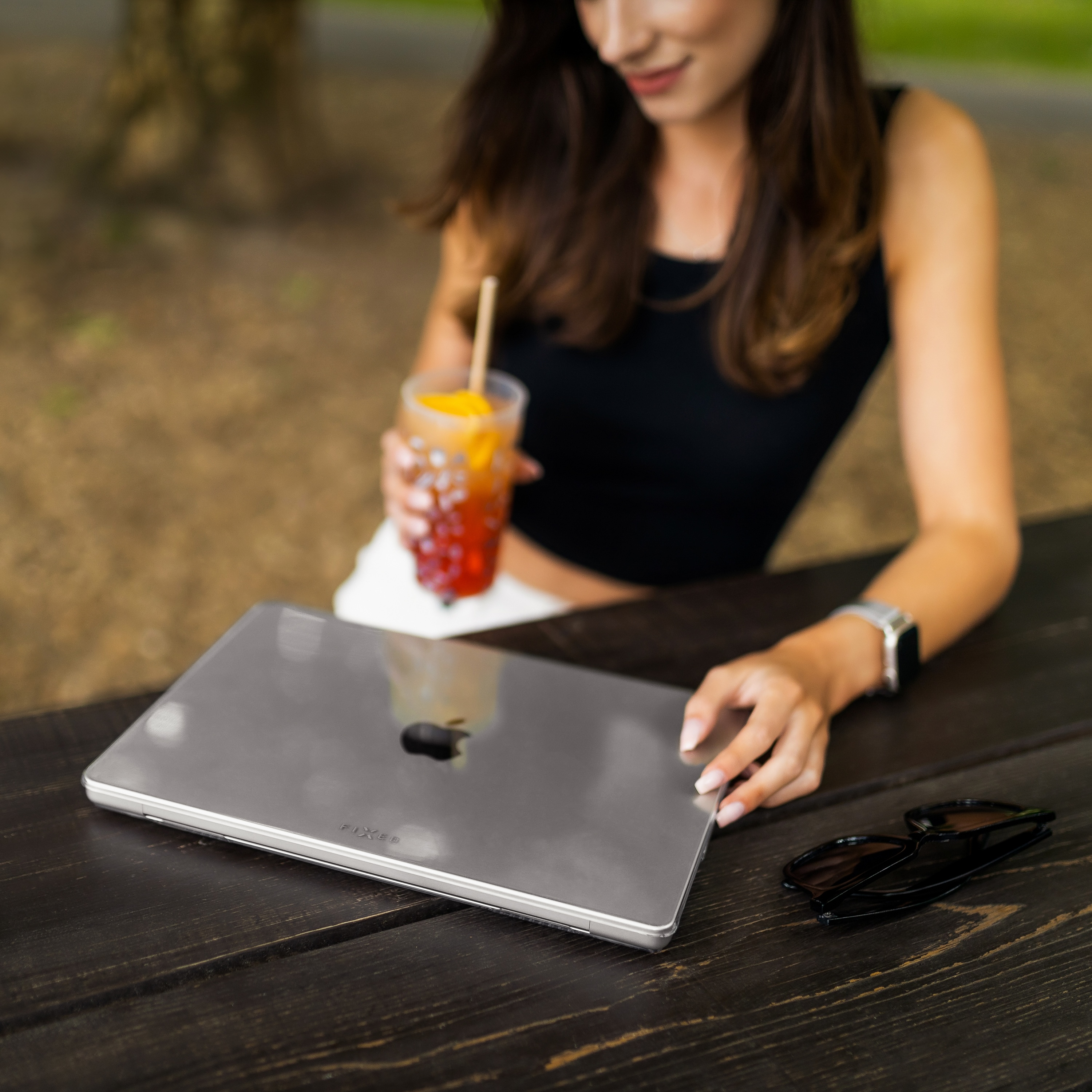 Frau mit Getränk und Laptop auf dem Tisch. Sonnenbrille liegt neben dem Laptop. Hintergrund ist unscharf.