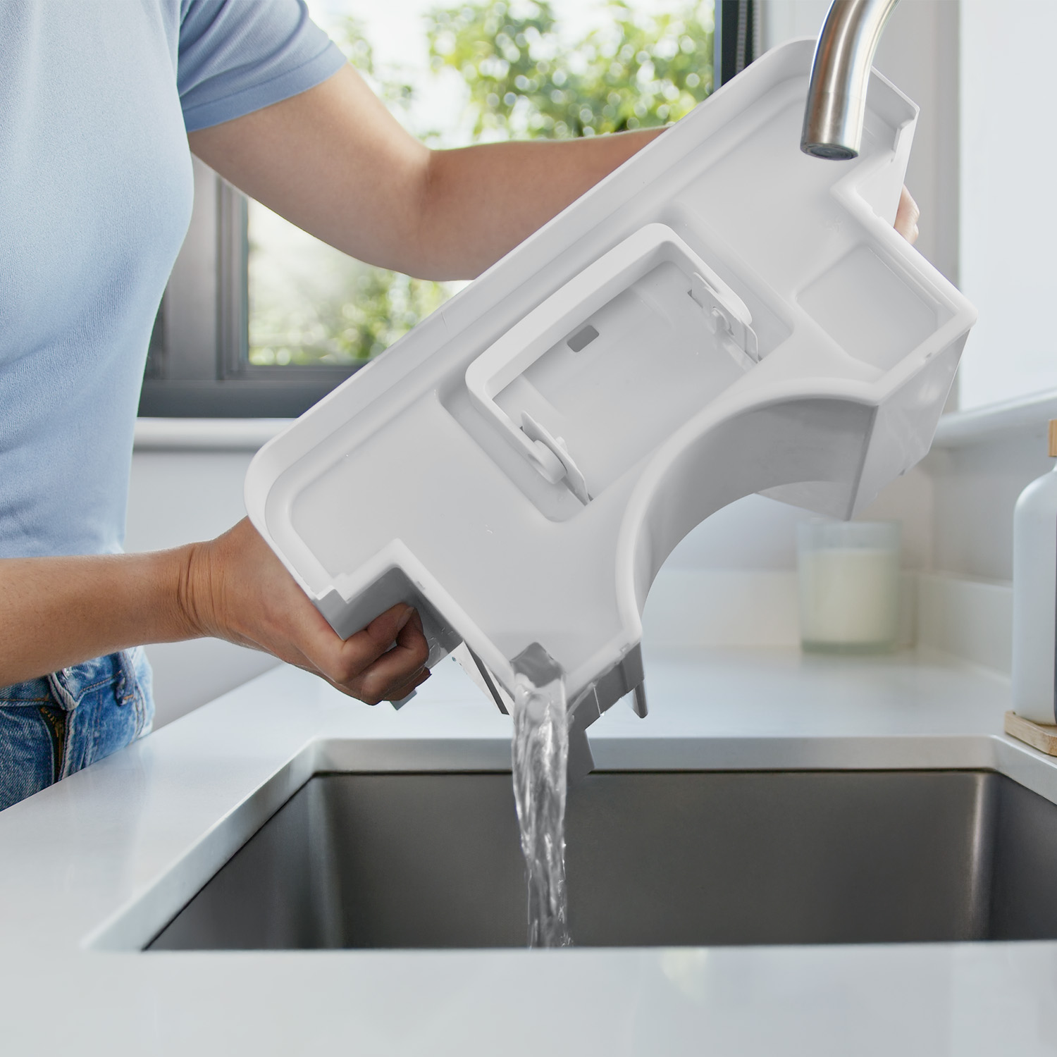 Eine Person gießt Wasser aus einem weißen Behälter in ein dunkles Waschbecken. Die Szene spielt in einer gut beleuchteten Küche.