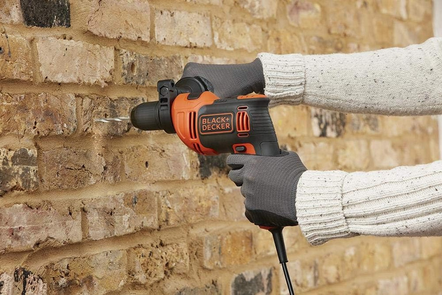 Una persona usa un trapano Black+Decker su un muro di mattoni. Indossa guanti e un maglione.