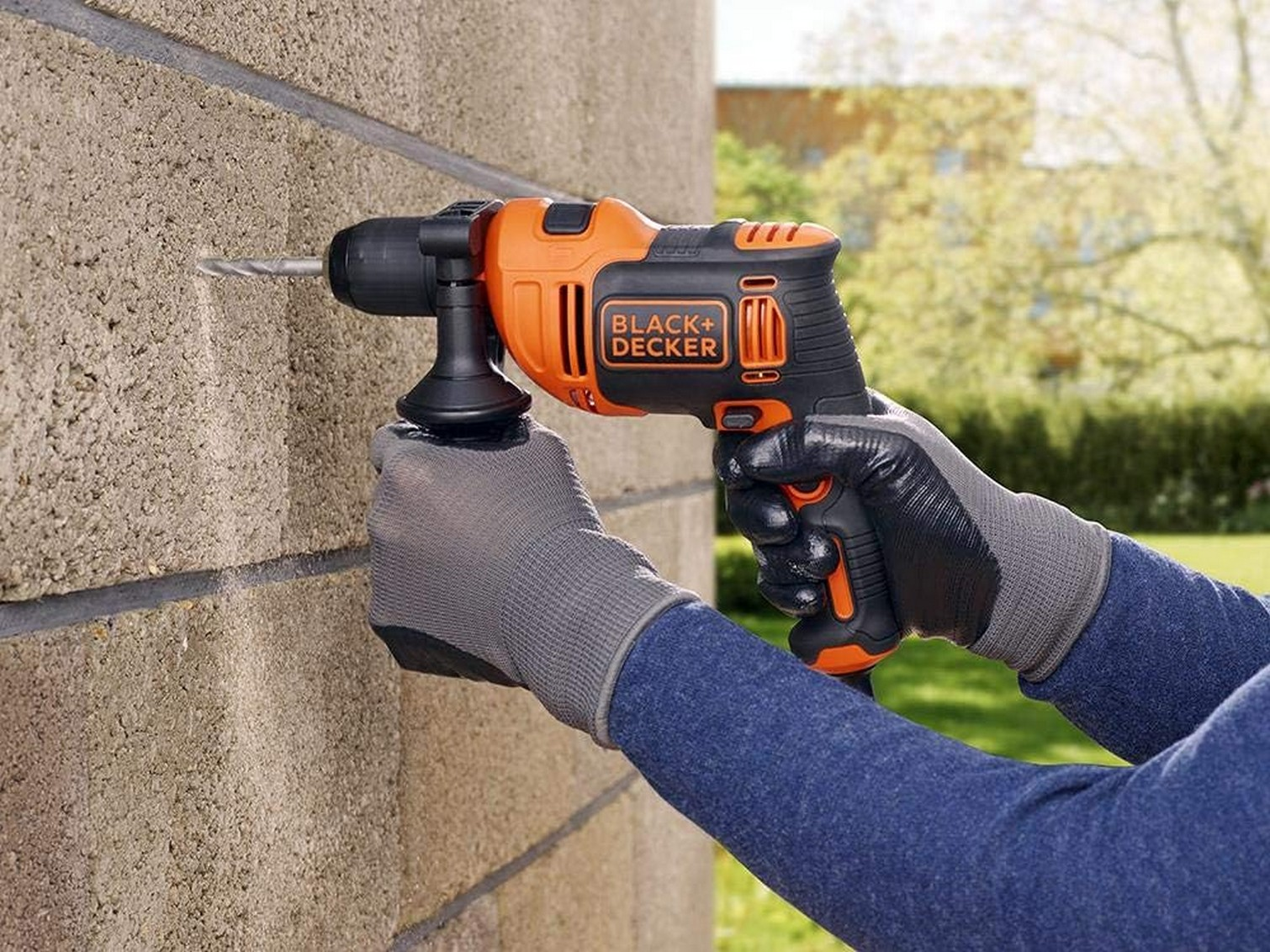 Una persona fora un muro di mattoni con un trapano Black+Decker arancione e nero. Indossa guanti e una camicia blu.