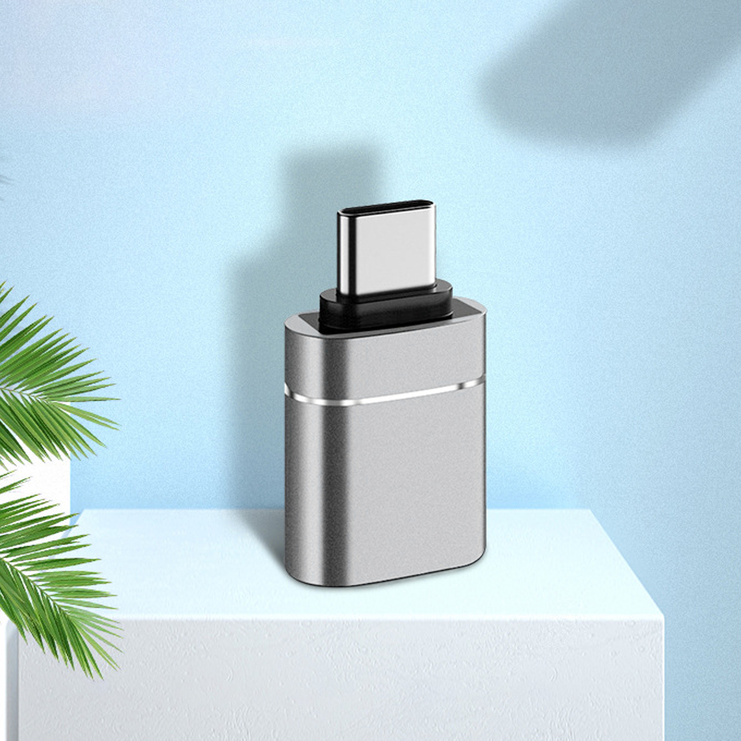 Srebrny adapter USB-C stoi na białej platformie na niebieskim tle. Zielone liście w lewym rogu.