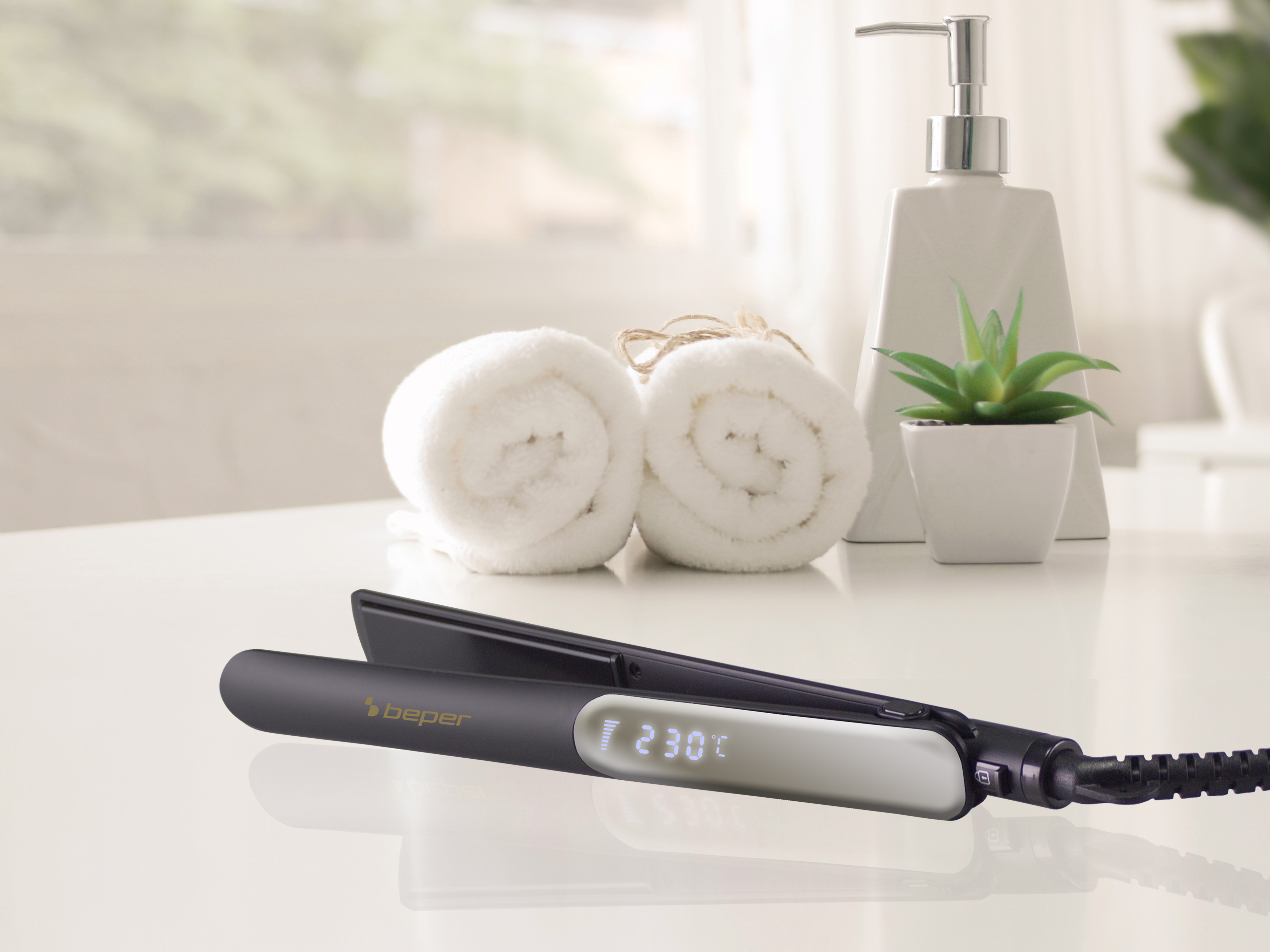 Una piastra per capelli con display digitale, due asciugamani bianchi arrotolati, un dosatore di sapone e una piccola pianta, su una superficie bianca.