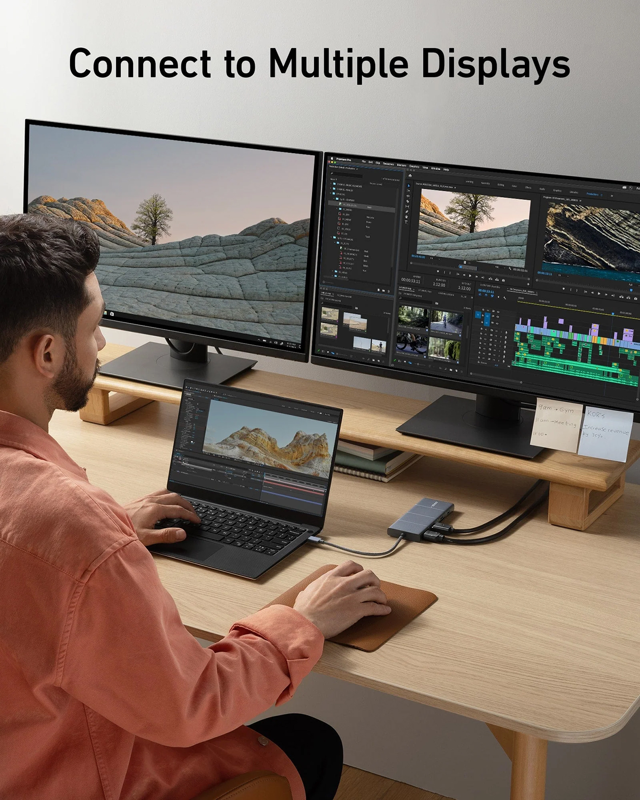 Un uomo usa un laptop collegato a due monitor. La scena mostra un software di editing video, una scrivania di legno e un hub.