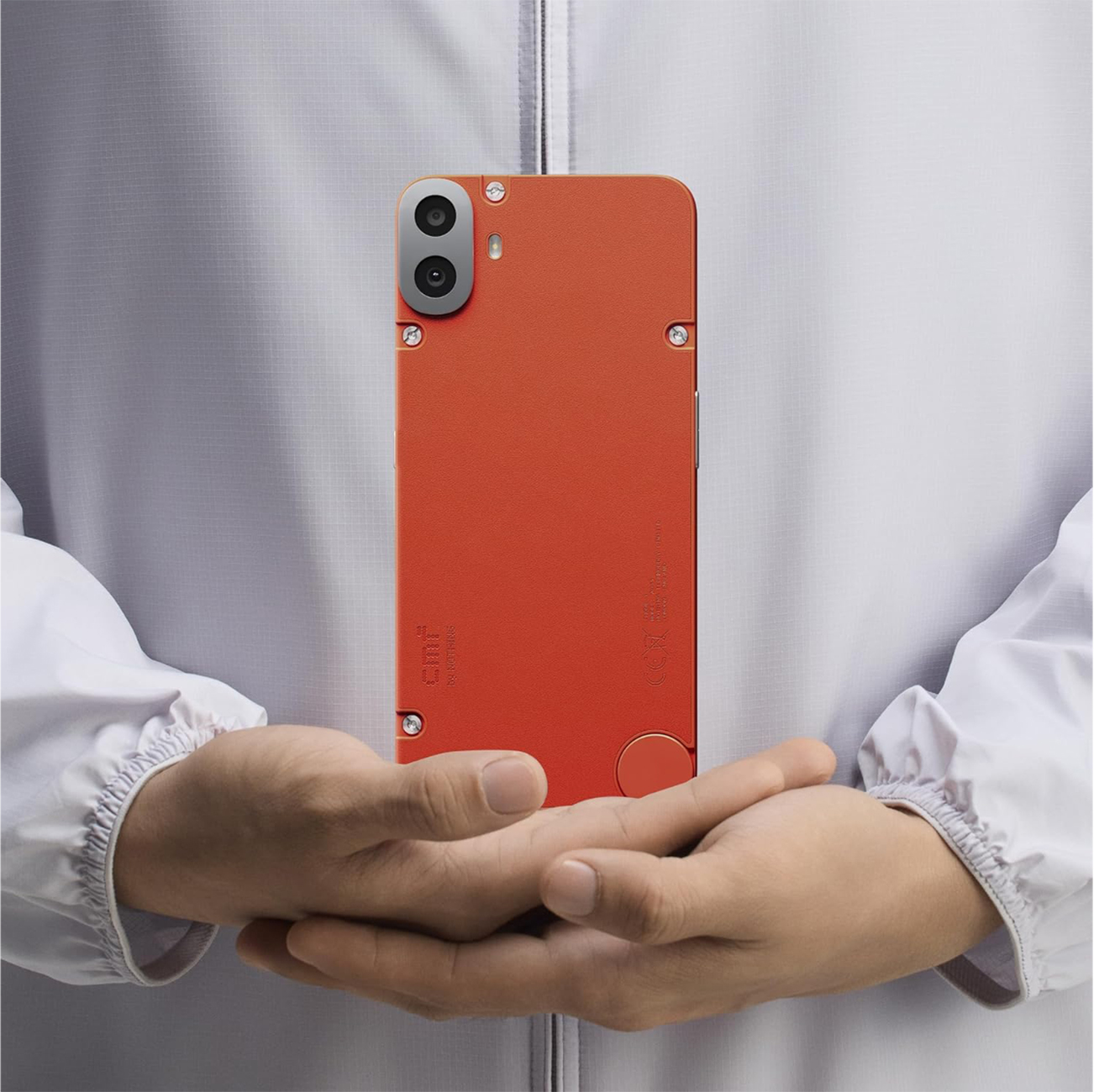 Eine Person hält ein orangefarbenes Telefon. Das Telefon hat eine Dual-Objektiv-Kamera und wird vor einer weißen Jacke gehalten.