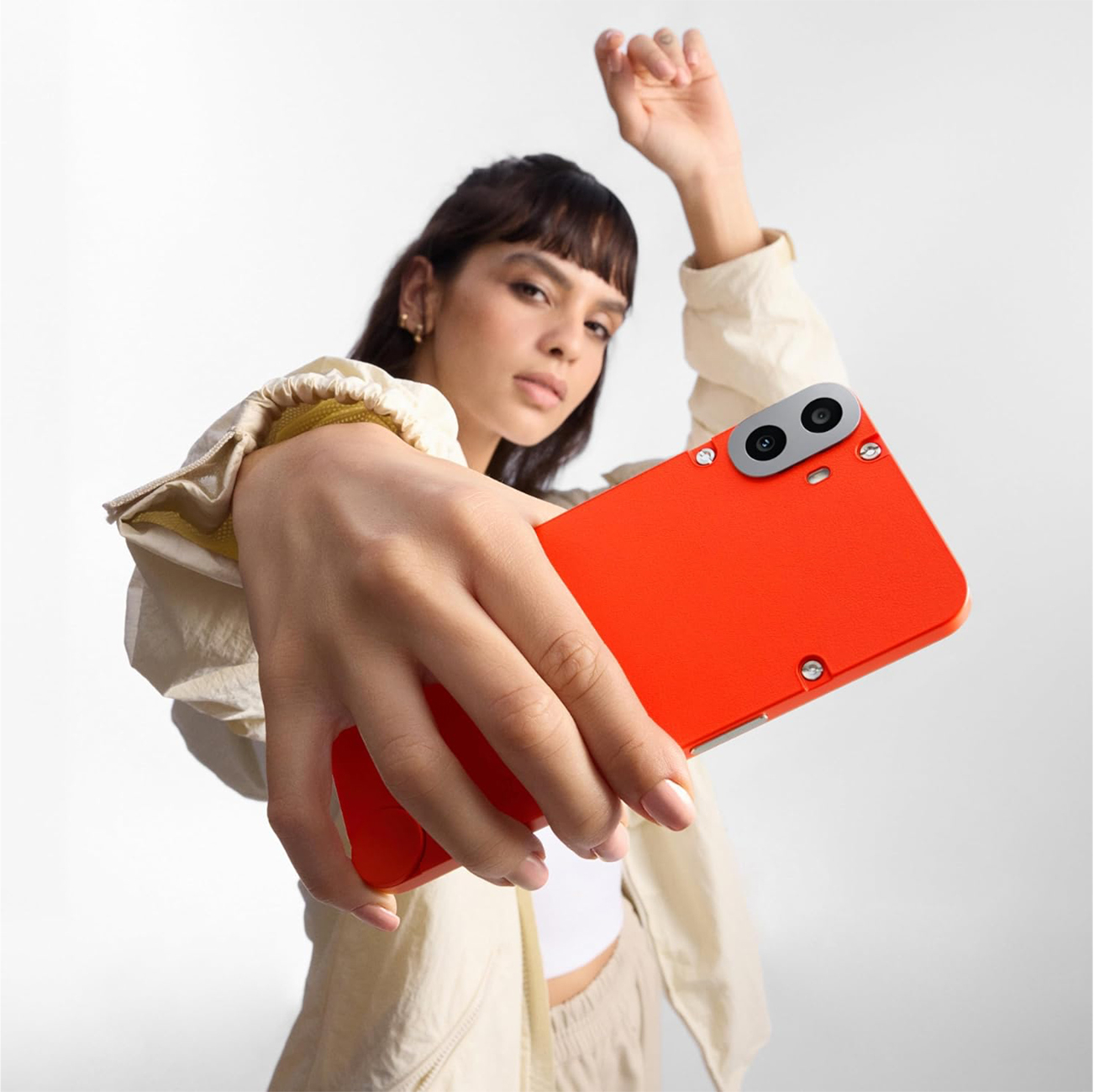 Eine Frau hält ein orangefarbenes Telefon und hebt den Arm. Sie trägt eine cremefarbene Jacke und ein weißes Oberteil. Der Hintergrund ist weiß.