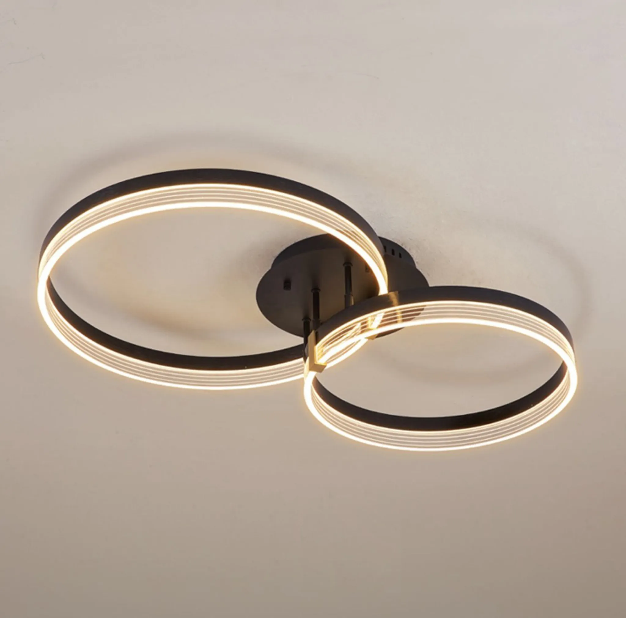 Eine moderne Deckenleuchte mit zwei sich kreuzenden, leuchtenden kreisförmigen Ringen, schwarzem Rahmen und weißer Decke.