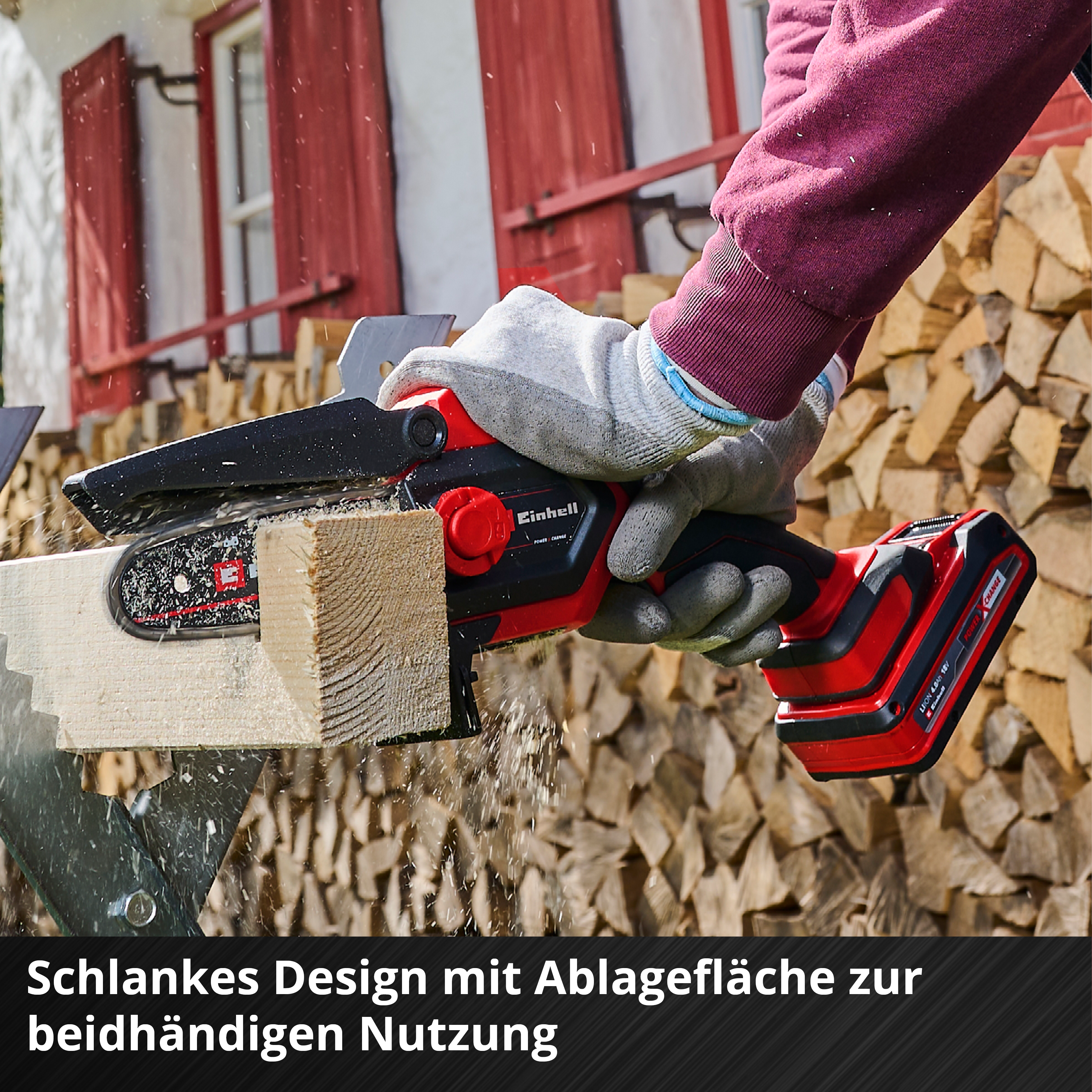 Eine Person schneidet mit einer rot-schwarzen Kettensäge Holz. Sägemehl fliegt. Hintergrund: gestapeltes Holz und rotes Gebäude.