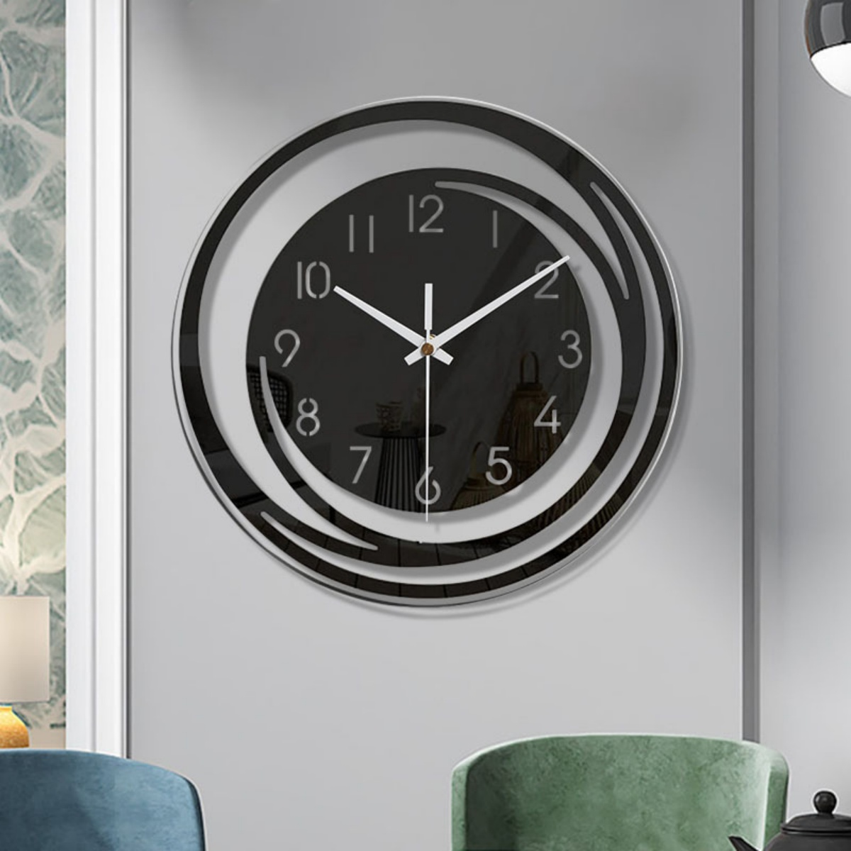 Eine moderne schwarz-silberne Wanduhr mit weißen Zeigern und Ziffern befindet sich mittig an einer grauen Wand. Im Vordergrund sind zwei grüne Stühle zu sehen.