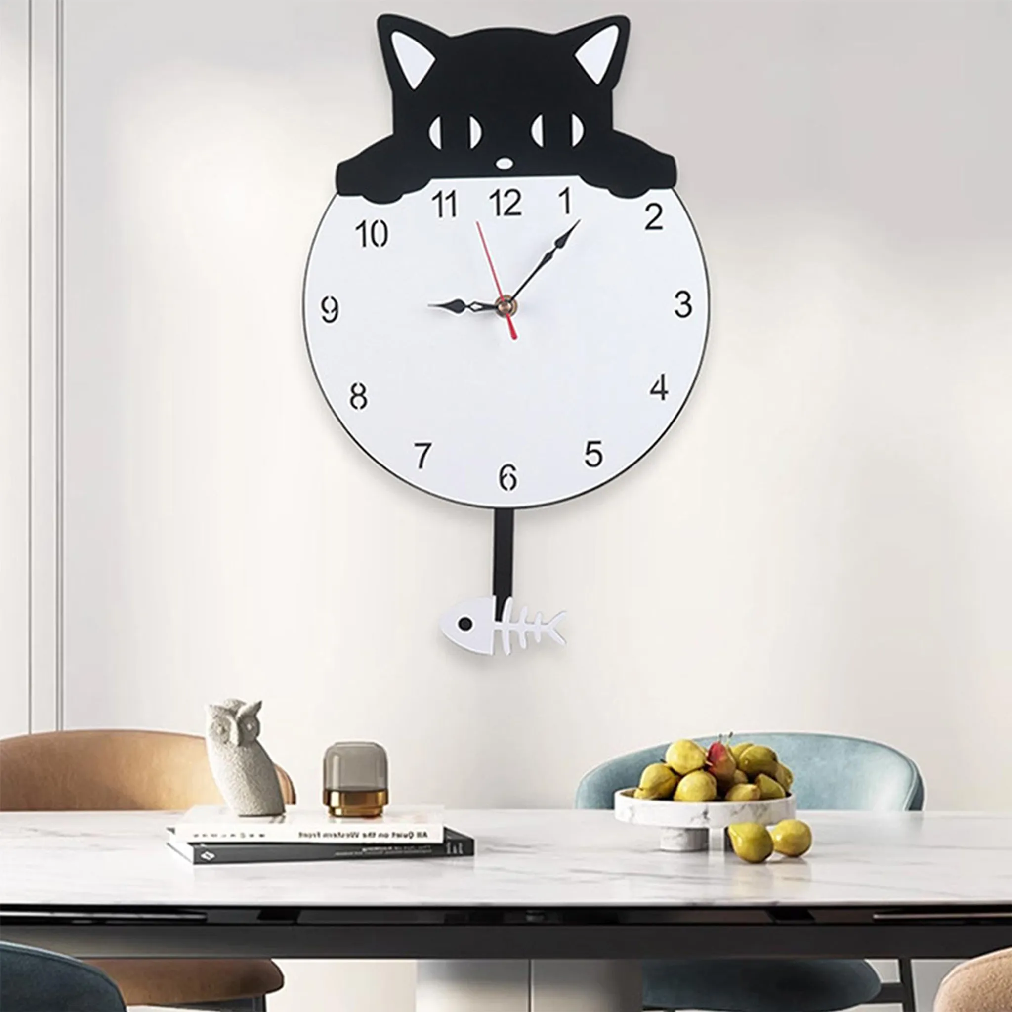 Eine katzenförmige Uhr hängt über einem Tisch. Eine Eulenstatue und Obst stehen auf dem Tisch. Stühle sind sichtbar.