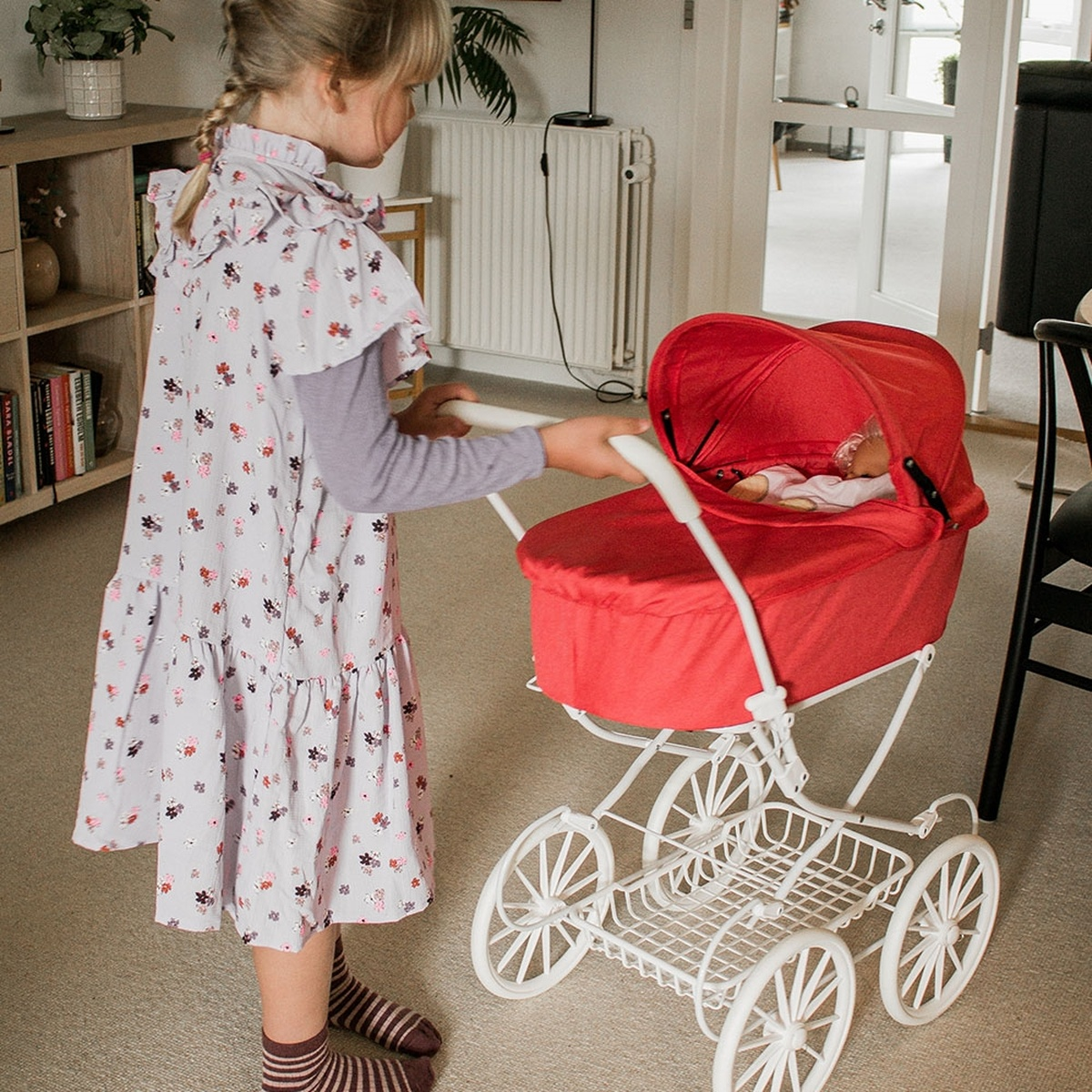 Ein Mädchen in einem geblümten Kleid schiebt einen roten Puppenwagen mit einer Puppe darin. Der Kinderwagen ist weiß mit weißen Rädern.