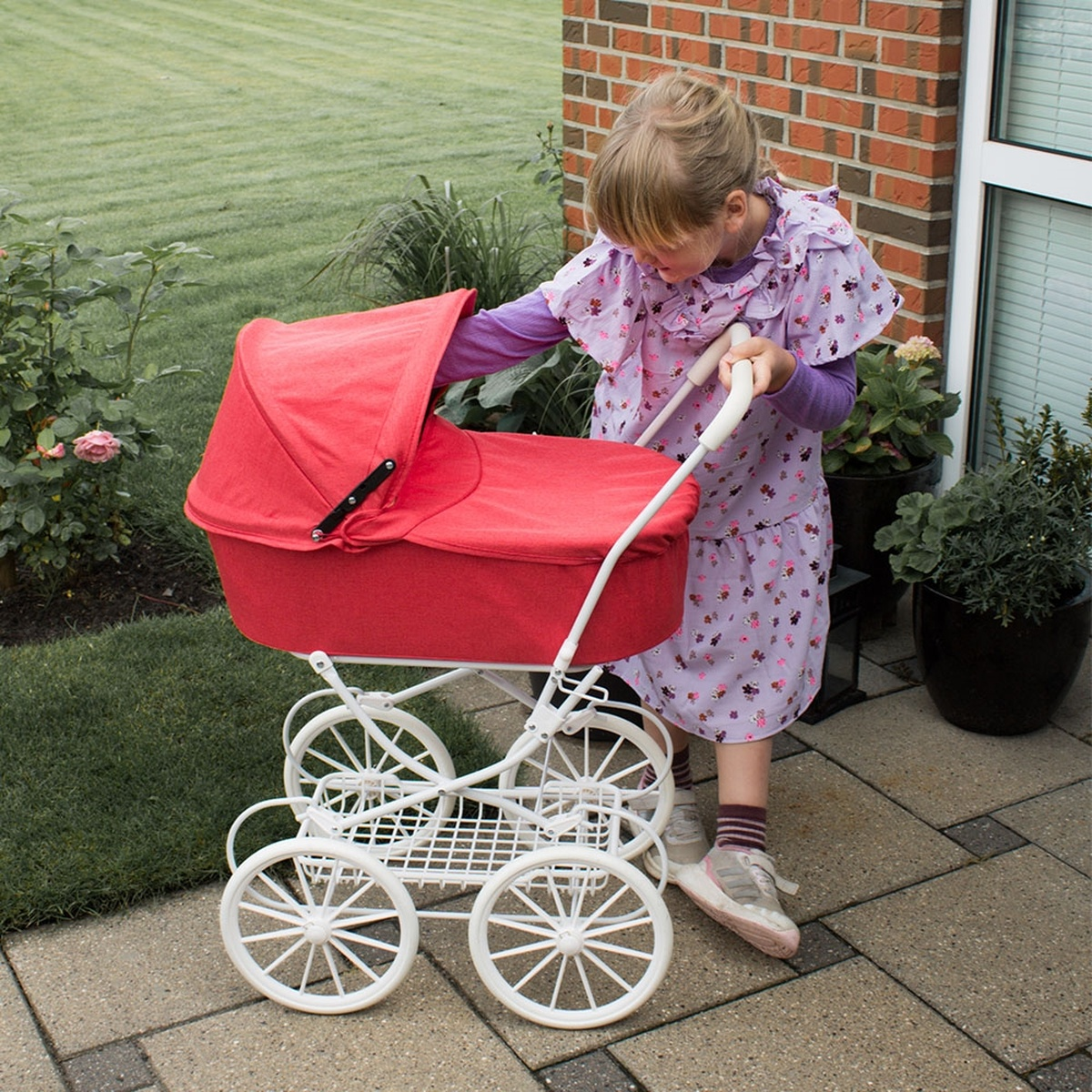 Ein Mädchen in einem geblümten Kleid schiebt einen roten Spielzeugkinderwagen. Der Wagen hat weiße Räder. Die Szene ist im Freien, in der Nähe einer Ziegelmauer und Pflanzen.