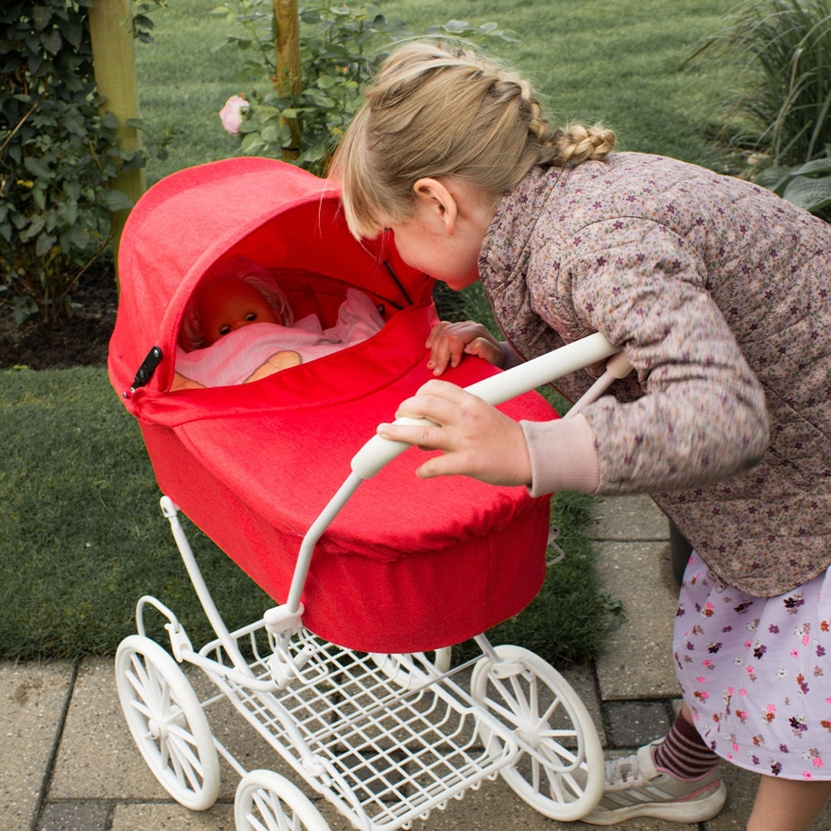 Ein Mädchen betrachtet eine Puppe in einem roten Kinderwagen. Der Kinderwagen ist weiß. Das Mädchen trägt eine Blumenjacke und einen Rock und steht auf einem Steinweg.