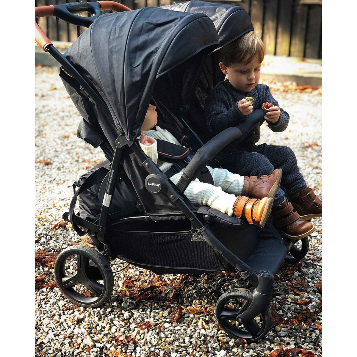Zwei Kinder in einem schwarzen Kinderwagen. Ein Kind hält ein kleines Objekt. Der Kinderwagen steht auf Kies und Herbstlaub.