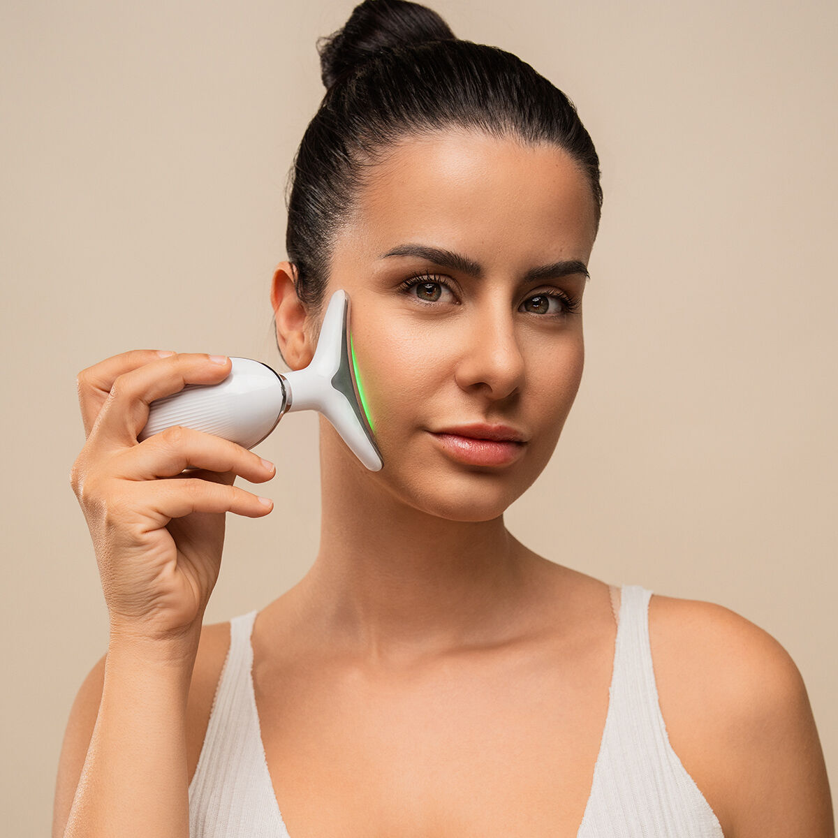 Una donna tiene un dispositivo facciale bianco sulla guancia. Il dispositivo ha una luce verde. Indossa un top bianco e i suoi capelli sono raccolti.