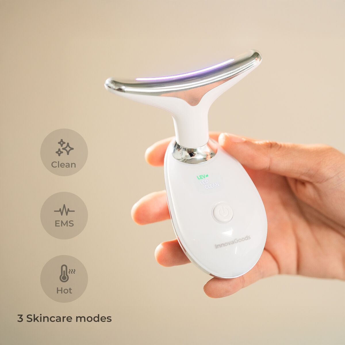 Un dispositivo bianco per la cura della pelle con testa argentata è tenuto in mano. Ha tre modalità: pulizia, EMS e caldo. Il logo InnovaGoods è visibile.
