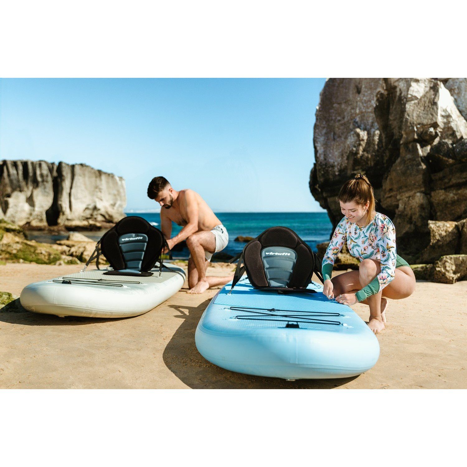 Zwei Personen bereiten Paddleboards an einem Sandstrand vor. Ein Mann und eine Frau knien. Im Hintergrund sind ein großer Felsen und blauer Himmel zu sehen.