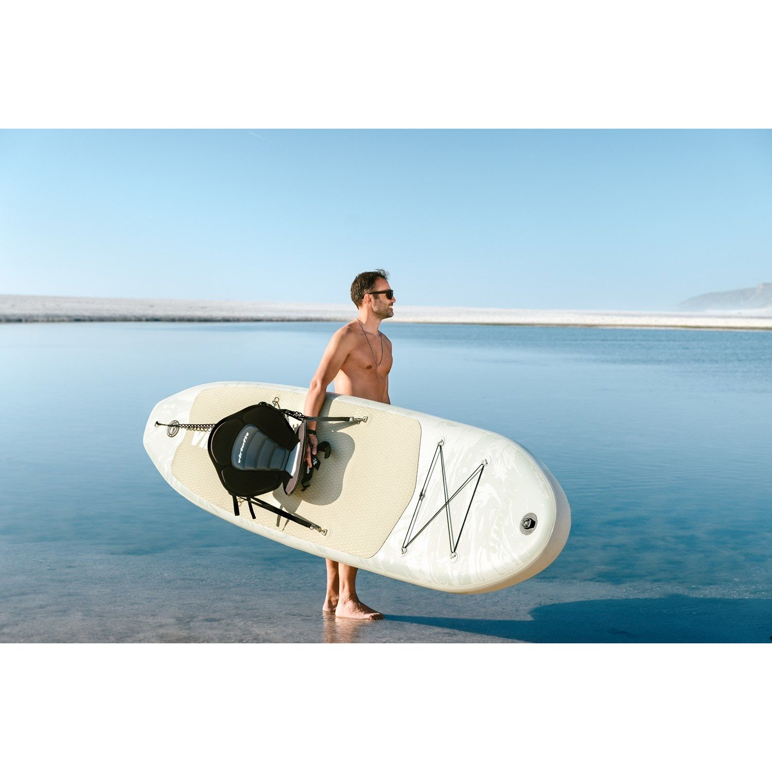 Ein Mann trägt ein Paddleboard und einen Sitz am Strand. Er ist ohne Hemd, trägt eine Sonnenbrille und steht im Wasser.