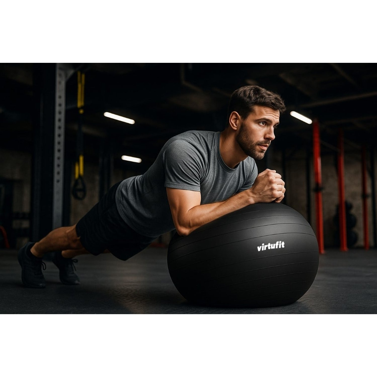 Ein Mann im Fitnessstudio macht eine Plank auf einem schwarzen Gymnastikball. Er trägt ein graues Hemd und schwarze Shorts. Im Hintergrund sind Fitnessgeräte zu sehen.