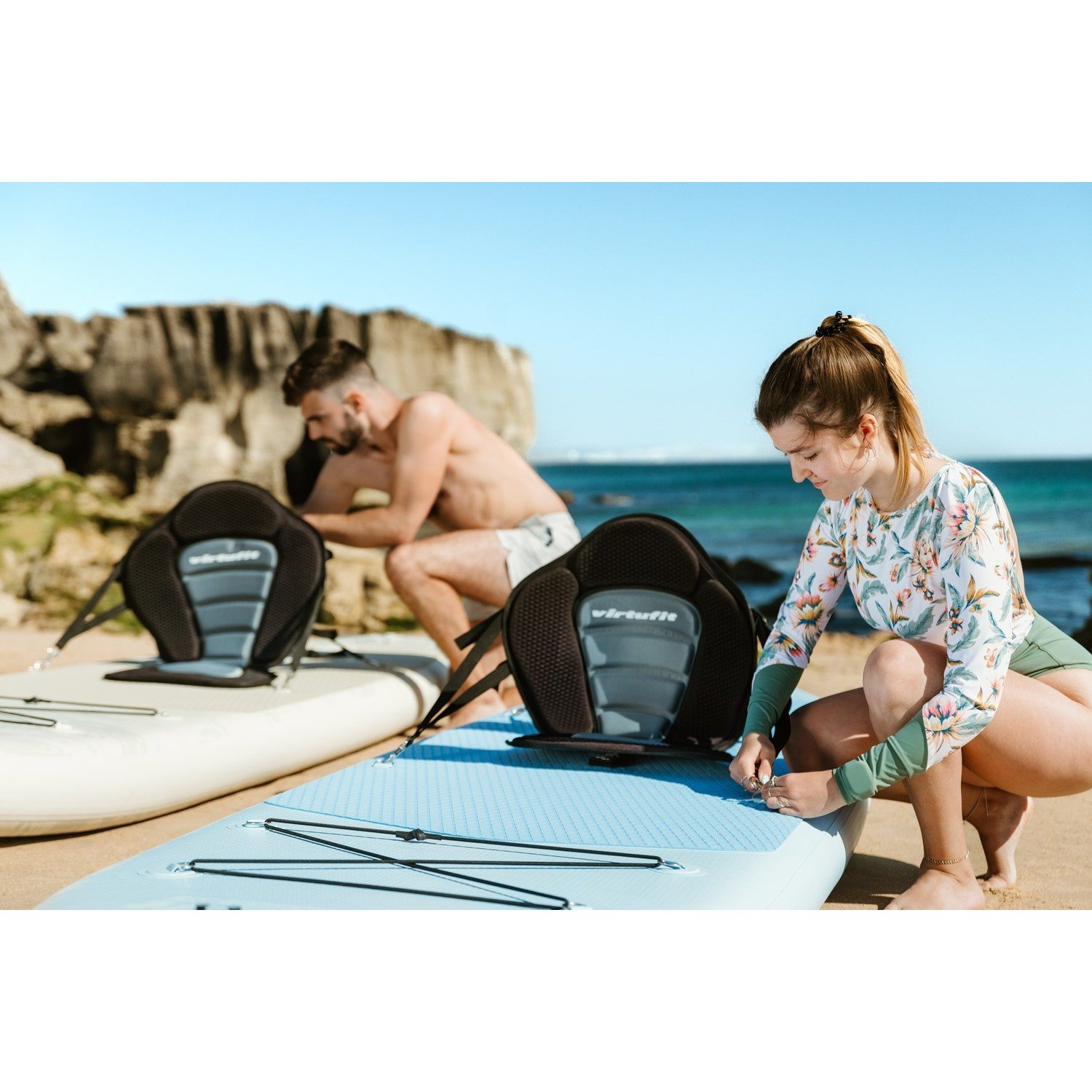 Eine Frau und ein Mann bereiten Paddleboards an einem Strand vor. Beide Paddleboards haben einen Sitz befestigt. Die Frau passt ihren Fuß an.