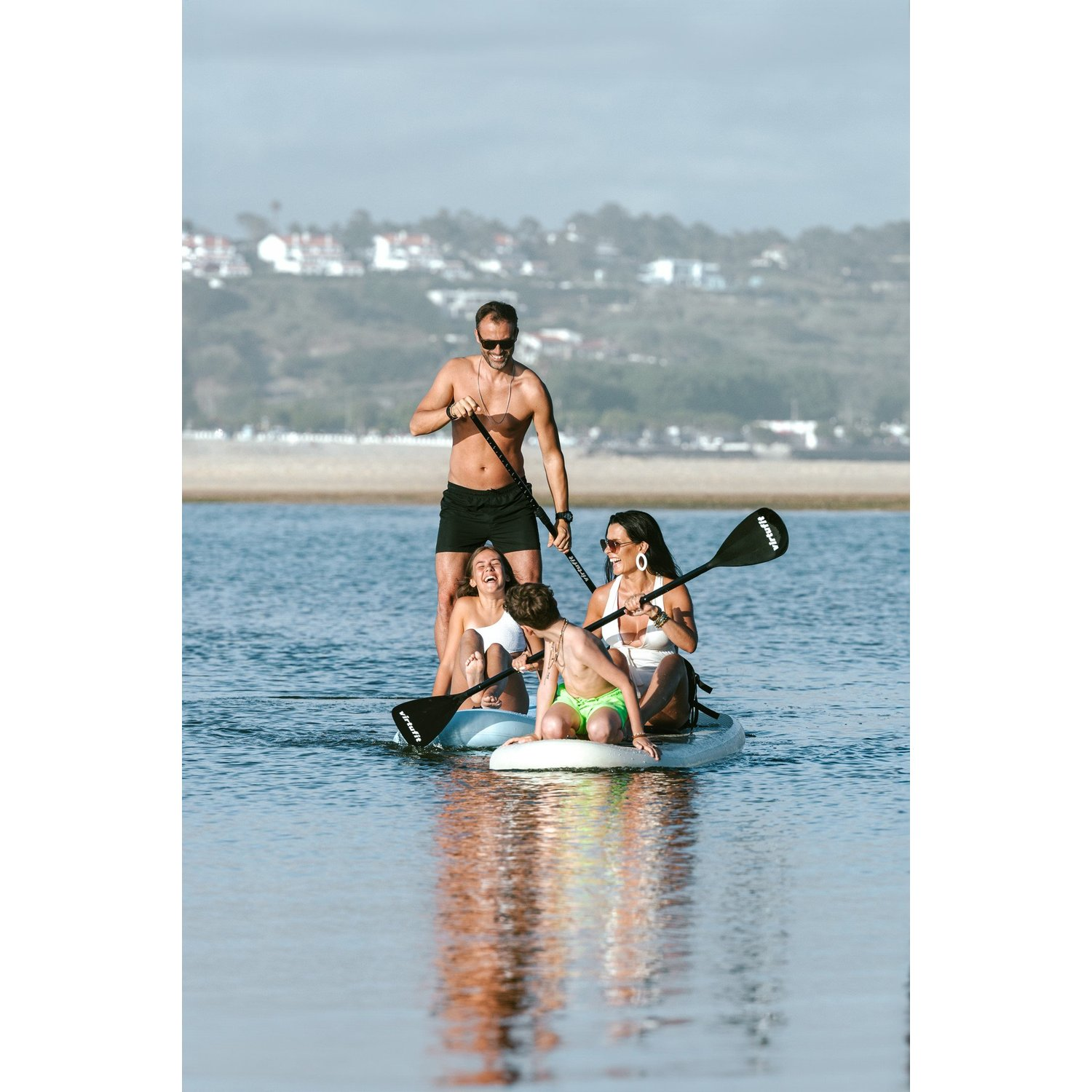 Familie auf Paddleboards. Mann paddelt, andere sitzen oder knien. Wasser reflektiert Himmel und Menschen. Häuser im Hintergrund.