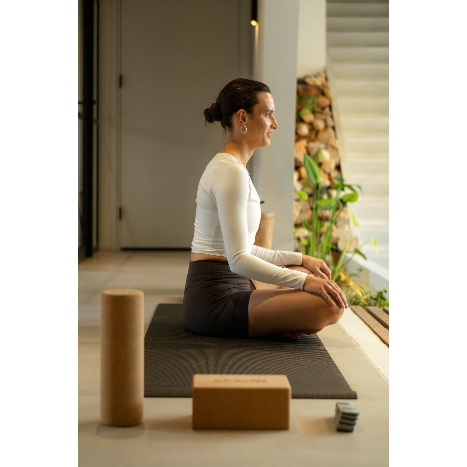 Frau in weißem Oberteil und Shorts sitzt auf einer Matte und meditiert. Yoga-Utensilien wie Blöcke und Rolle sind vor ihr.
