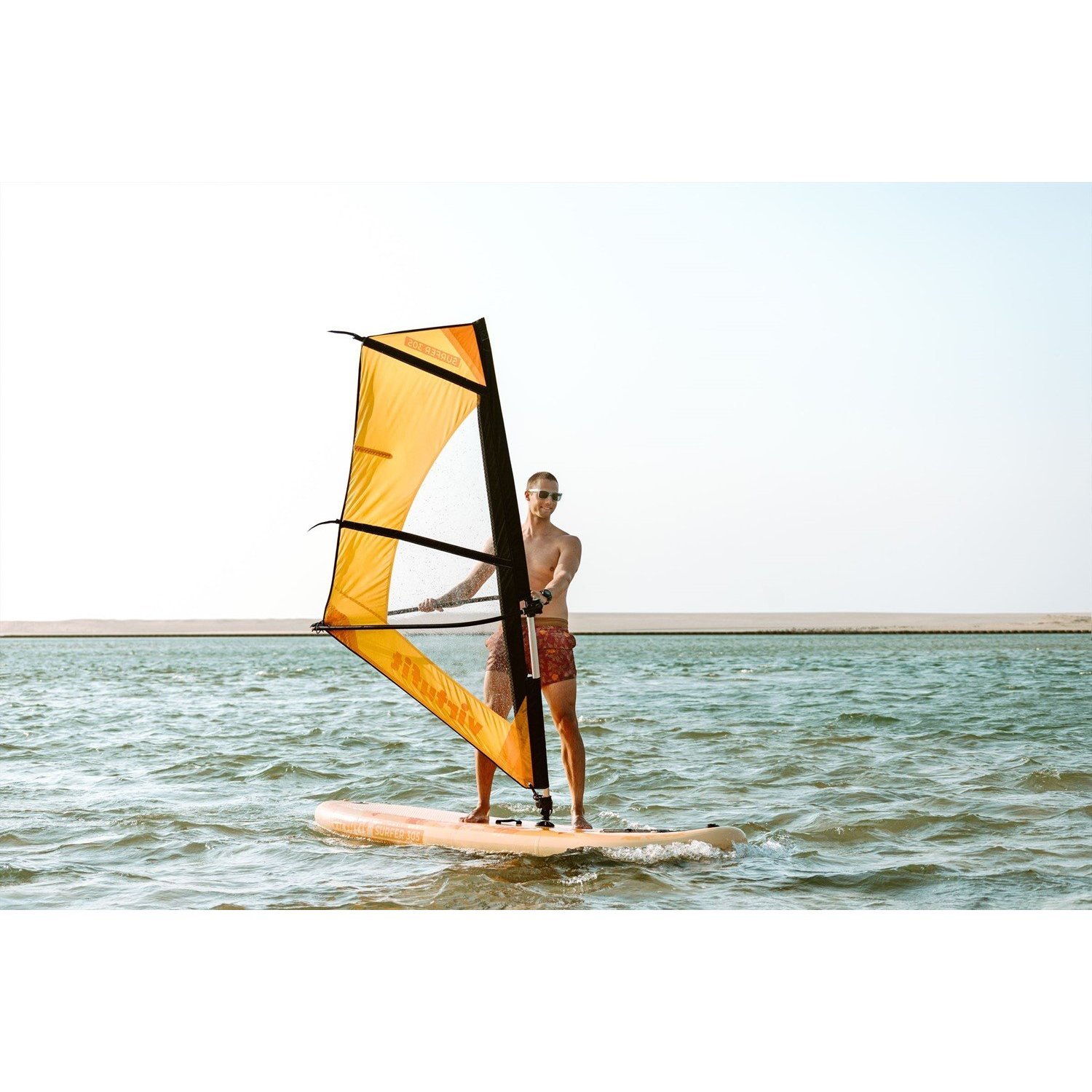 Ein Mann beim Windsurfen auf einem See. Er ist ohne Hemd und trägt eine Sonnenbrille und Shorts. Das Segel ist gelb und schwarz.