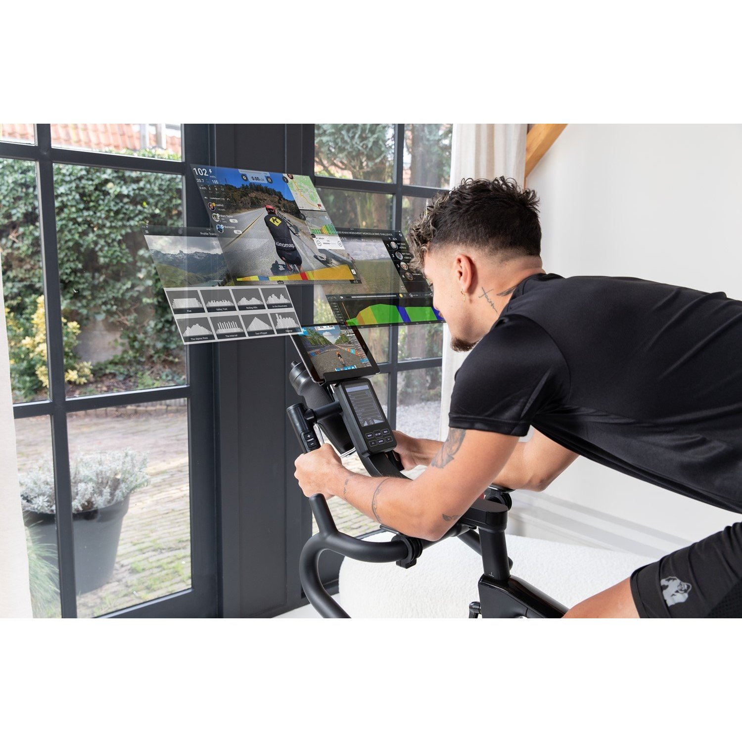 Ein Mann auf einem Heimtrainer betrachtet Bildschirme mit Fahrraddaten. Im Hintergrund ein Fenster mit Gartenblick.