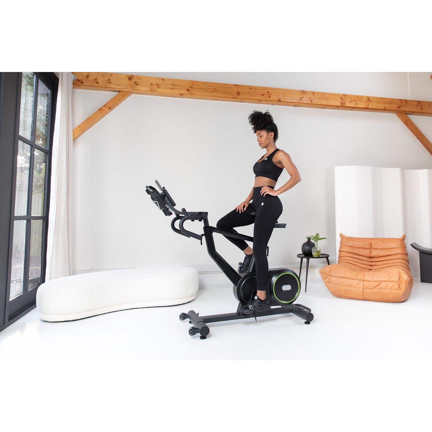 Eine Frau in schwarzer Sportkleidung auf einem Heimtrainer. Der Raum hat weiße Wände und einen weißen Boden. Es gibt eine weiße, flauschige Bank.