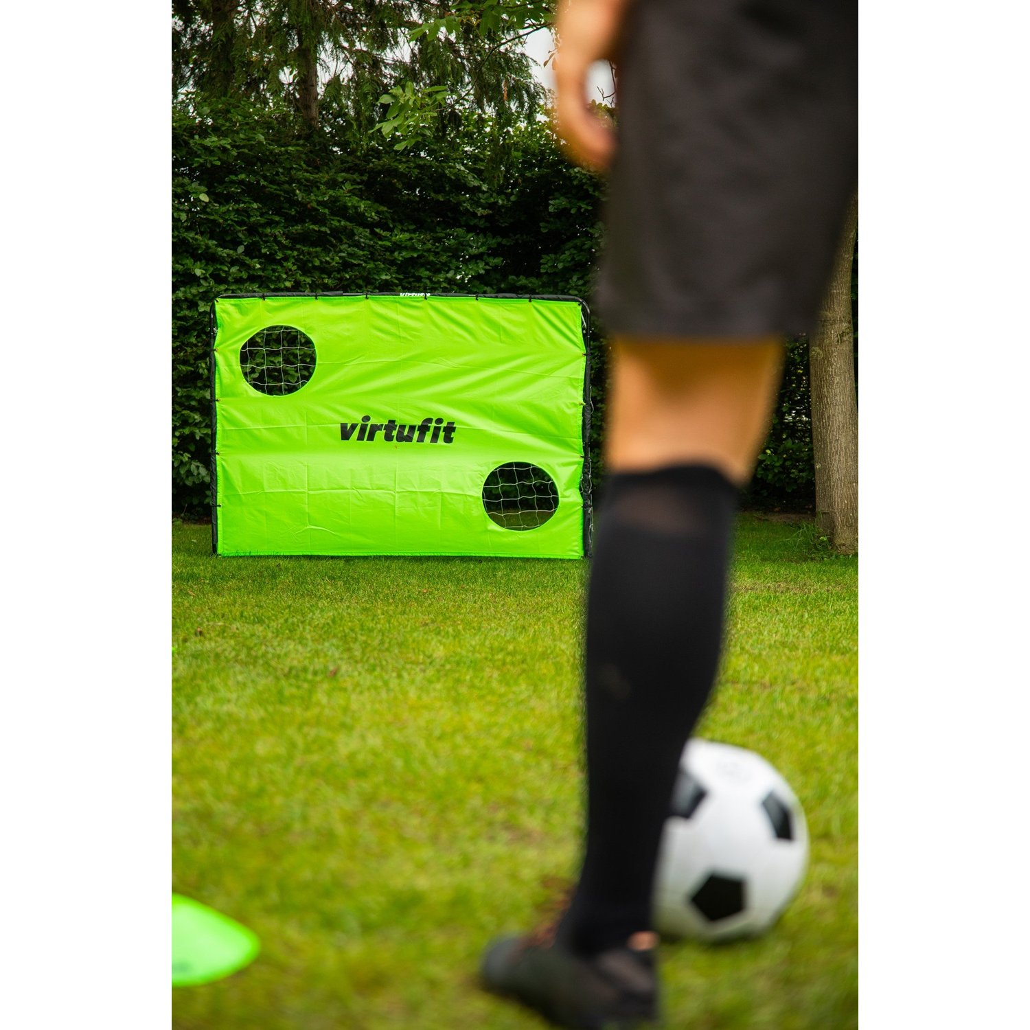 Un calciatore calcia un pallone verso una porta verde con due buchi. Sfondo: prato verde e cespugli.