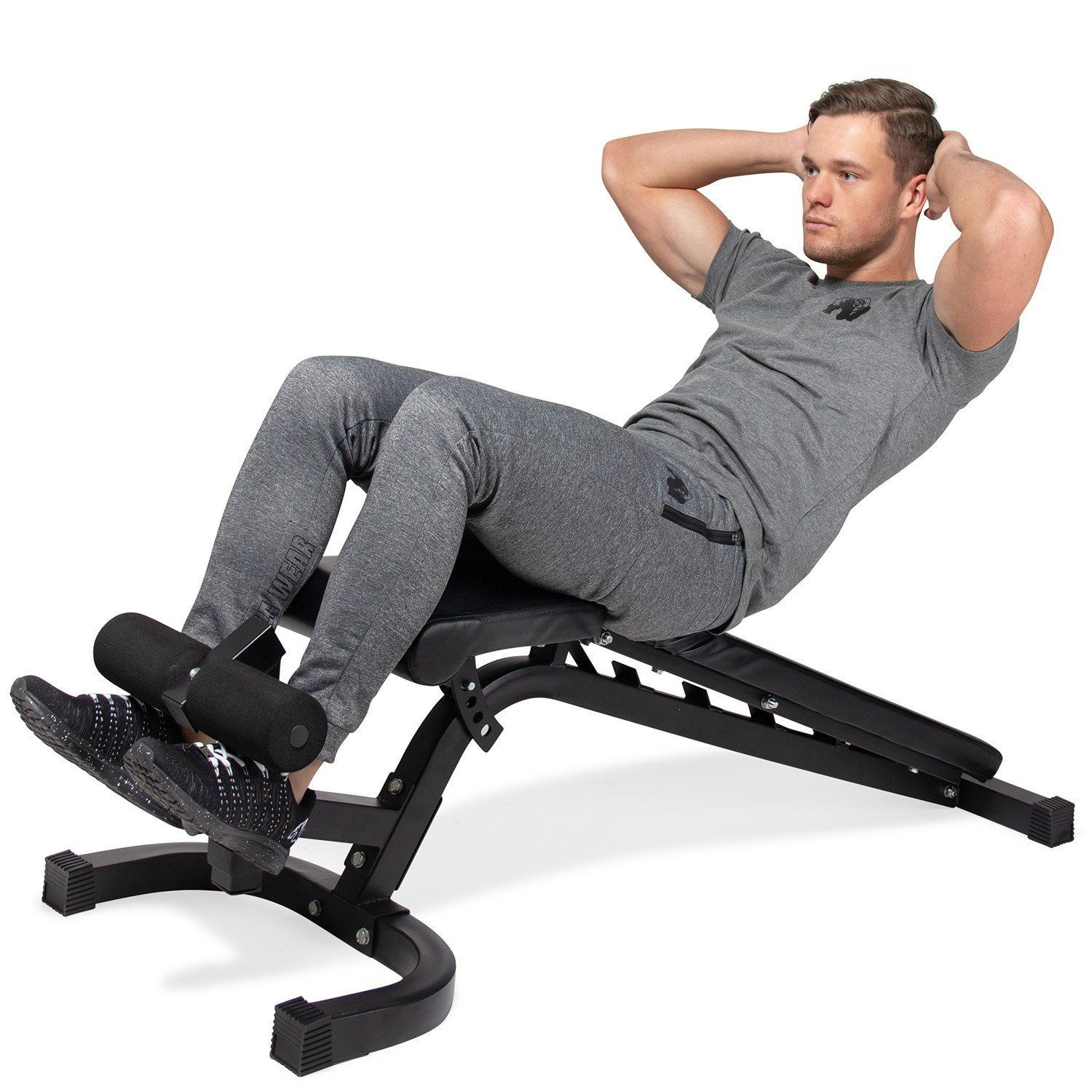 Ein Mann in grauer Jogginghose und einem Hemd macht Sit-ups auf einer schwarzen Trainingsbank. Seine Hände sind hinter seinem Kopf.