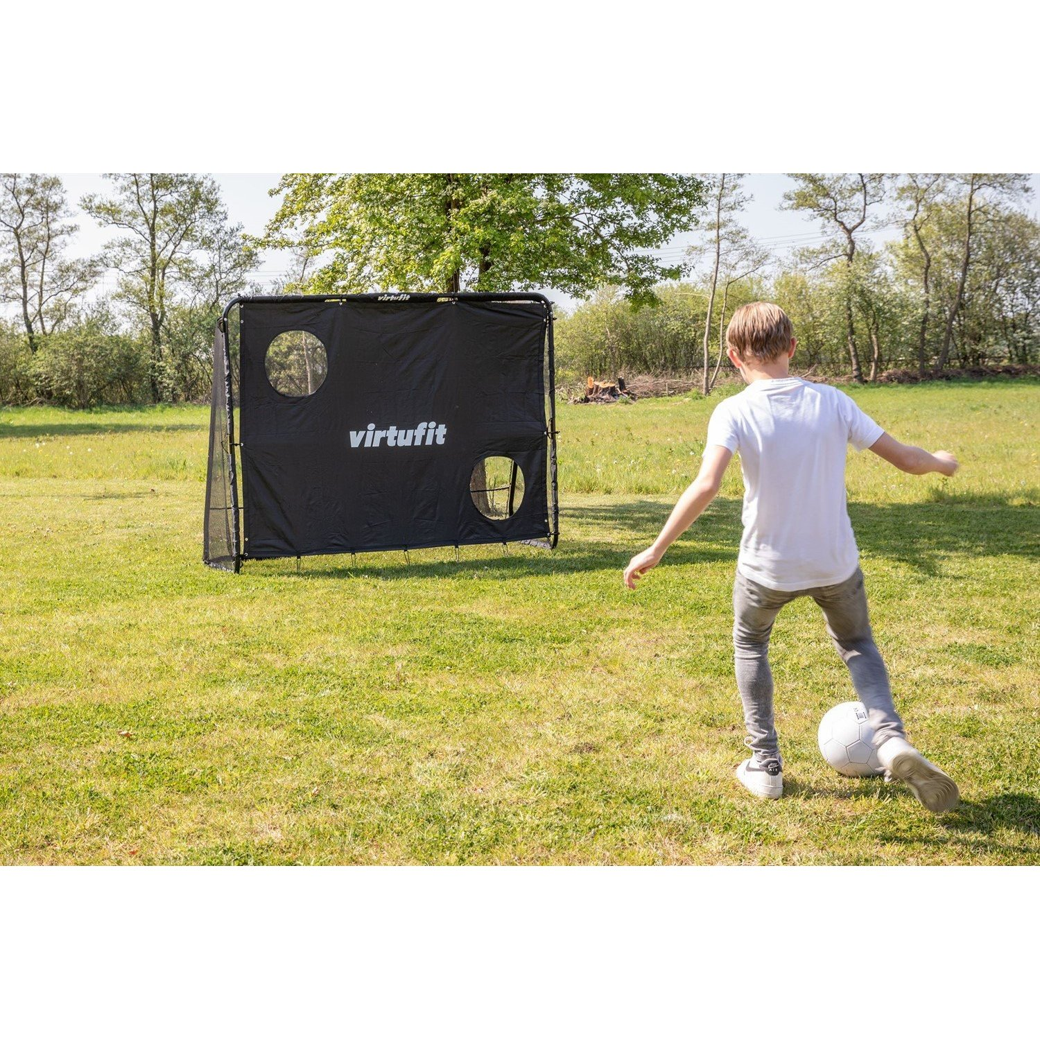 Un ragazzo calcia un pallone da calcio verso una porta nera con buchi su un campo erboso. Gli alberi sono sullo sfondo.