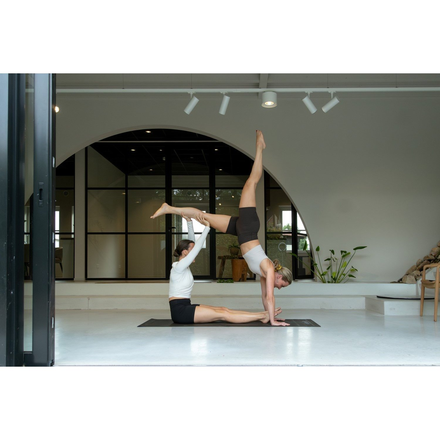 Zwei Frauen beim Yoga. Eine steht kopfüber und stützt die andere. Sie sind in einem weißen Studio mit offener Tür.