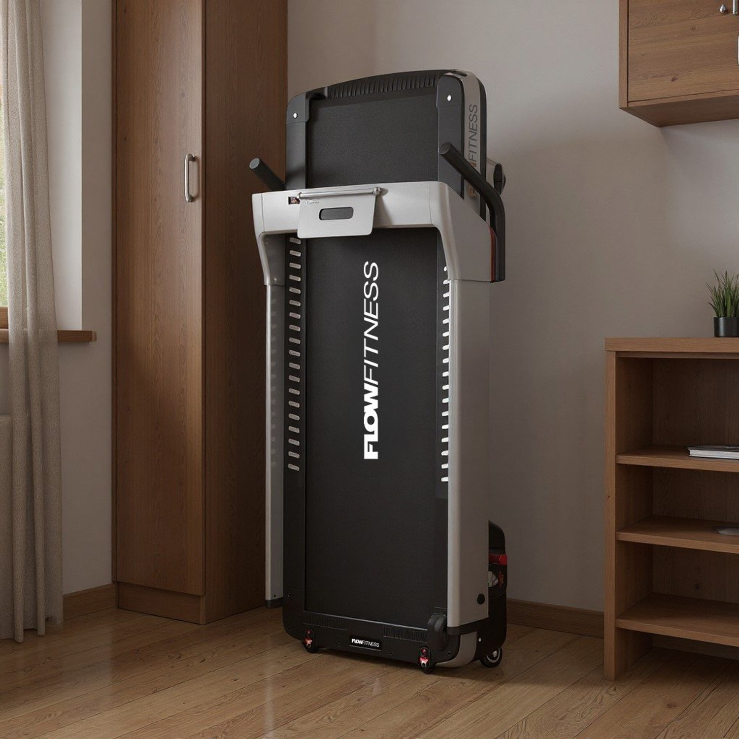 Ein zusammengeklapptes Laufband steht in einem Raum. Es ist schwarz und grau, mit dem Schriftzug FLOW FITNESS. Es gibt einen Holzschrank und Regale.