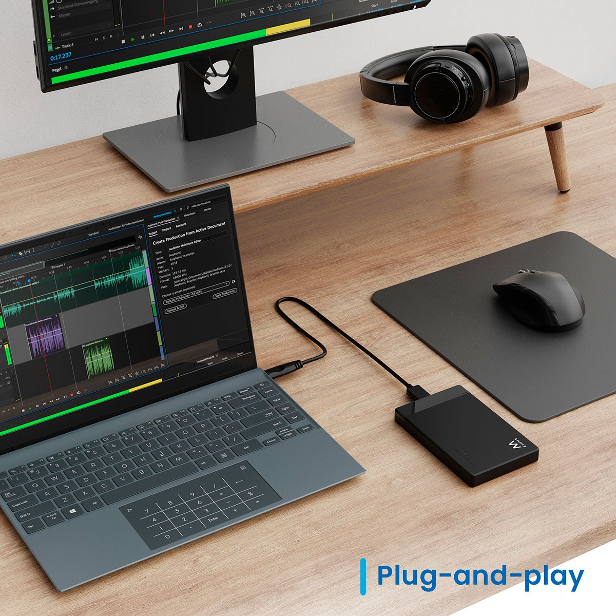 Un portátil, un monitor, unos auriculares y un disco duro externo están sobre un escritorio de madera. El portátil muestra una interfaz de edición de audio.