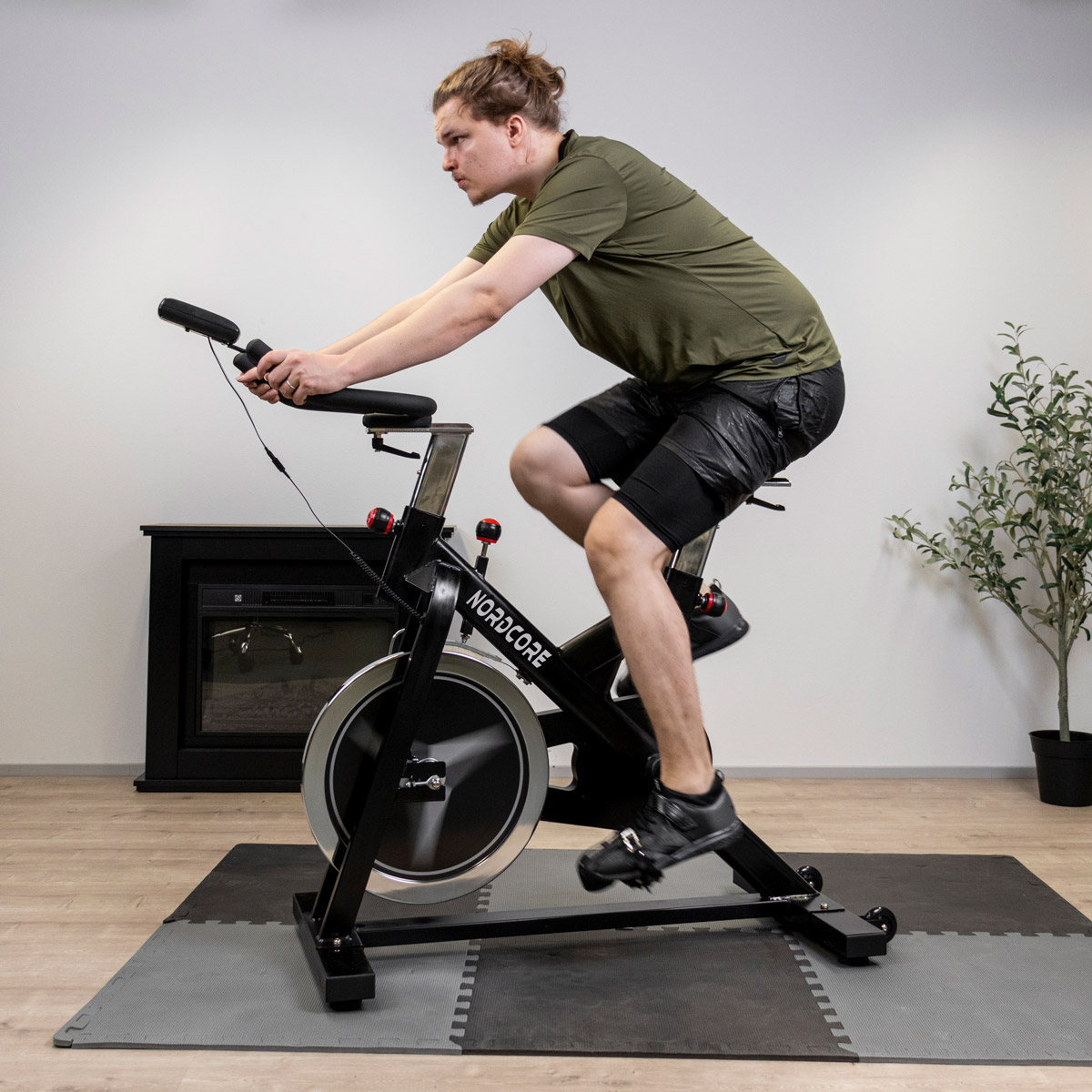 Mężczyzna jedzie na rowerze stacjonarnym. Ma na sobie zieloną koszulkę i czarne szorty. Rower jest czarny, ze srebrnym kołem.