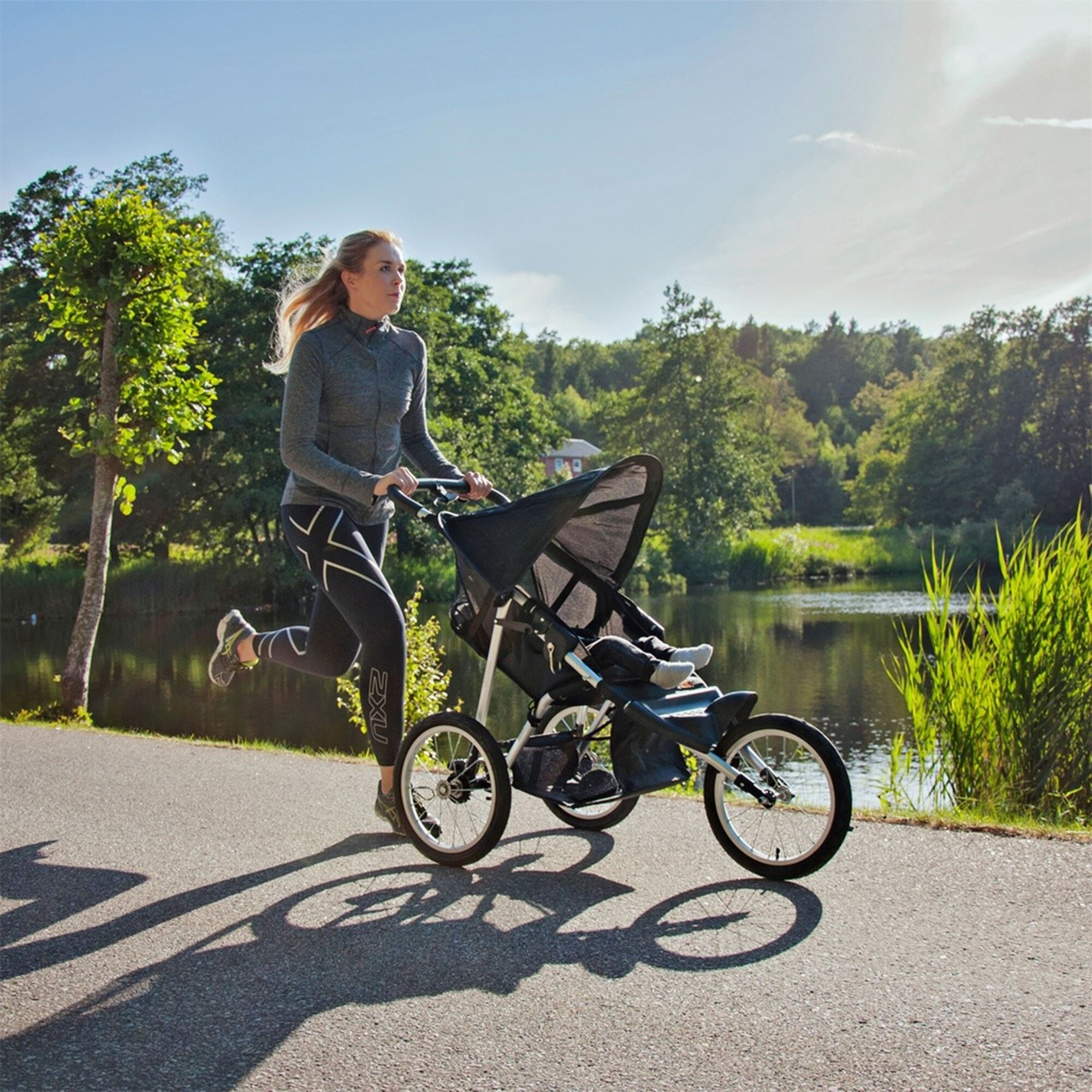 Frau rennt mit einem Kinderwagen auf einem Weg am See. Bäume und grünes Gras sind im Hintergrund. Sonniger Tag.
